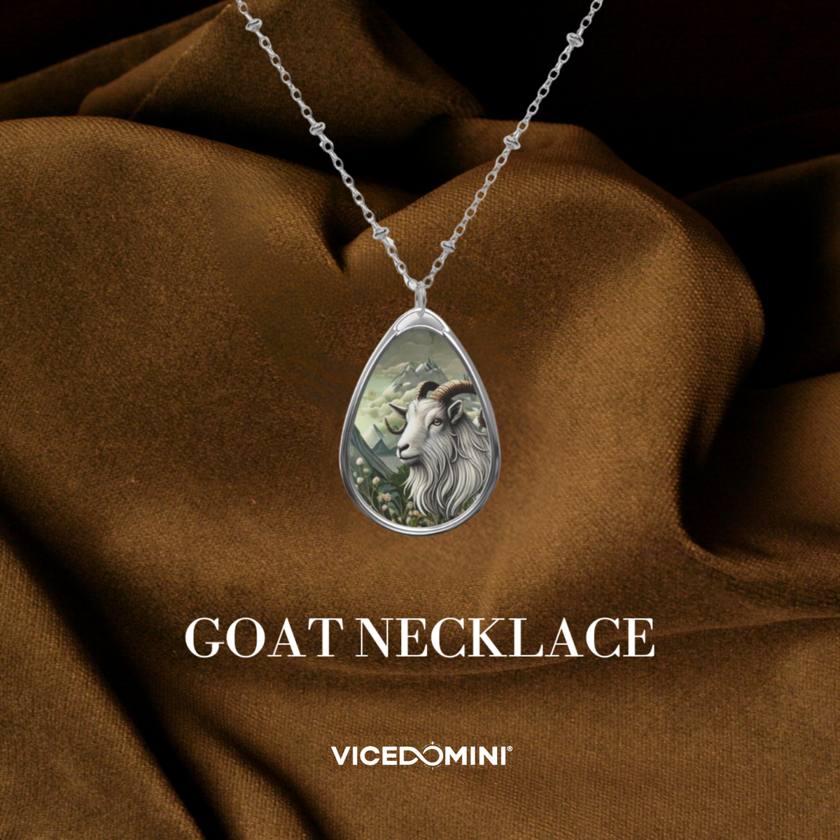 A teardrop-shaped silver pendant necklace featuring an illustrated goat head, displayed on draped brown fabric with the text 'GOAT NECKLACE' and 'VICEDOMINI' below.