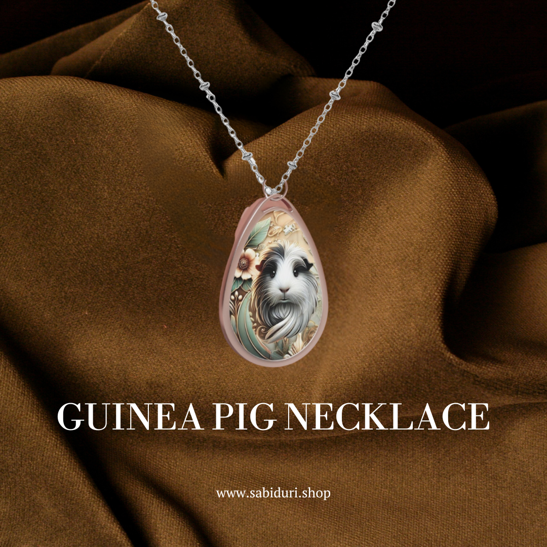 A close-up of the guinea pig necklace, showing the same intricate pendant design, resting on a luxurious brown fabric background. The text "GUINEA PIG NECKLACE" is written in white at the bottom, along with the URL "www.sabiduri.shop."