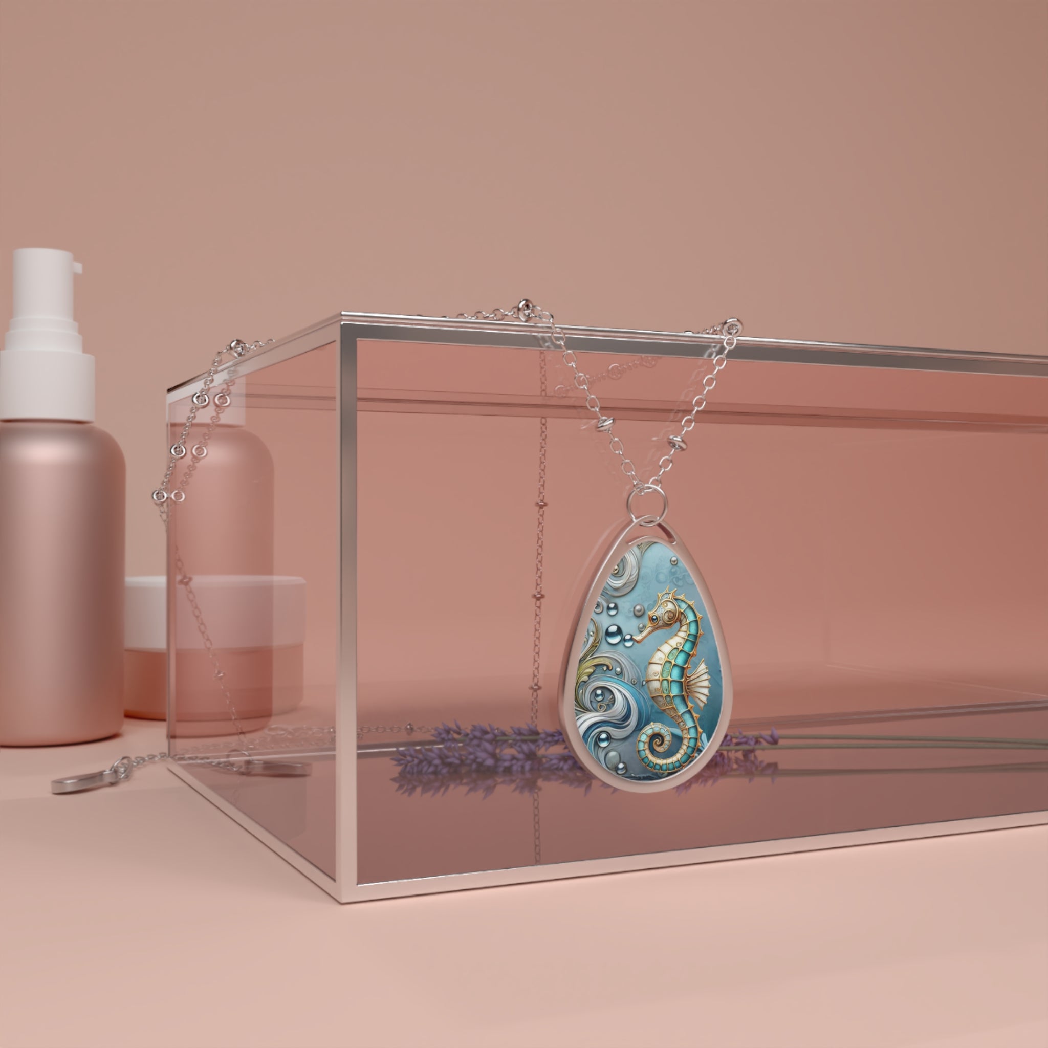 A teardrop pendant necklace with a seahorse design in blue and gold is displayed on a glass box. The necklace is draped elegantly over the edge of the box, which is set against a soft pink background with matching cosmetic bottles.