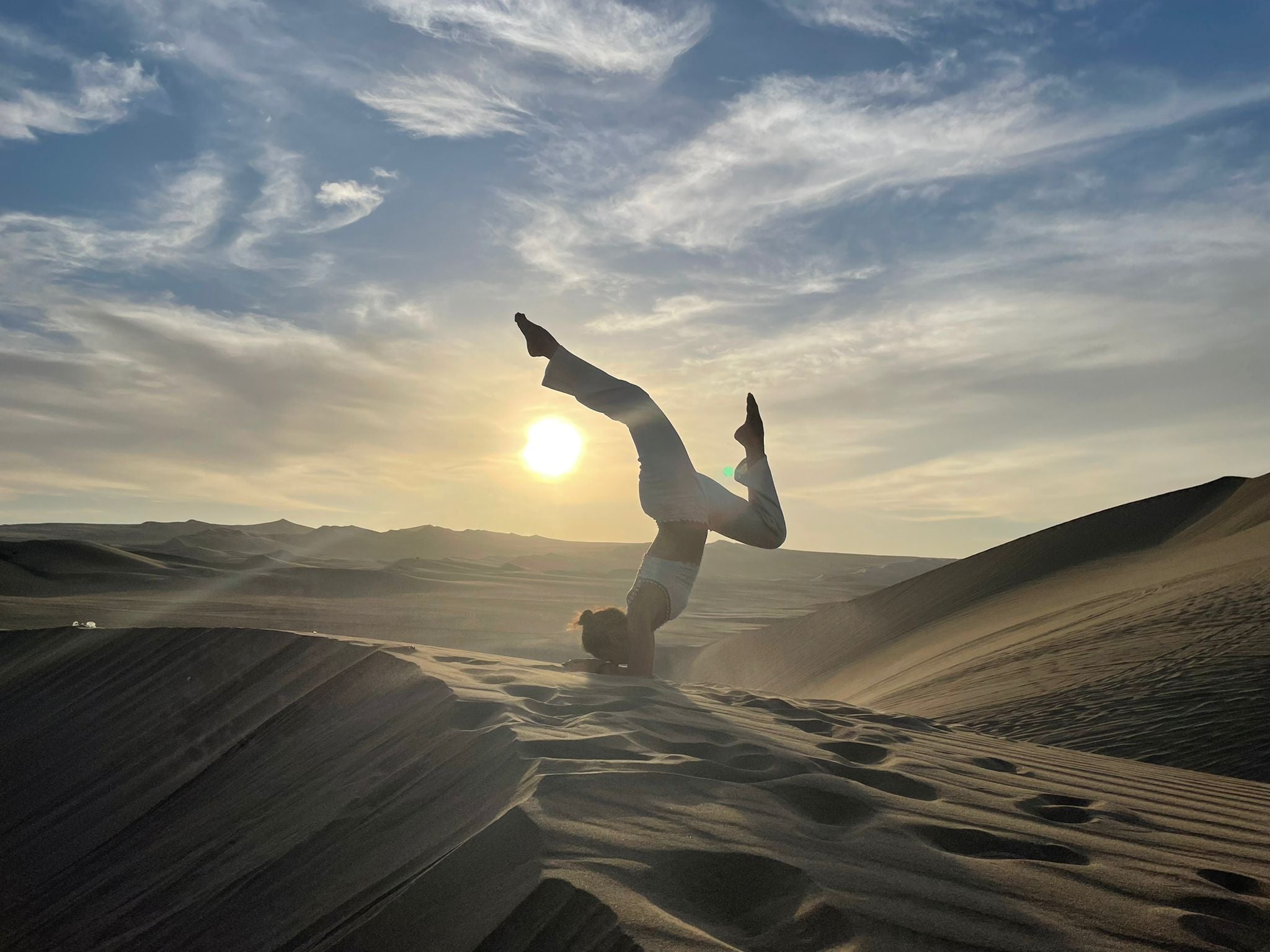 A silhouette of a person performing a one-handed handstand on a desert dune against a beautiful sunset sky, with the sun casting a warm glow over the rippled sand texture.