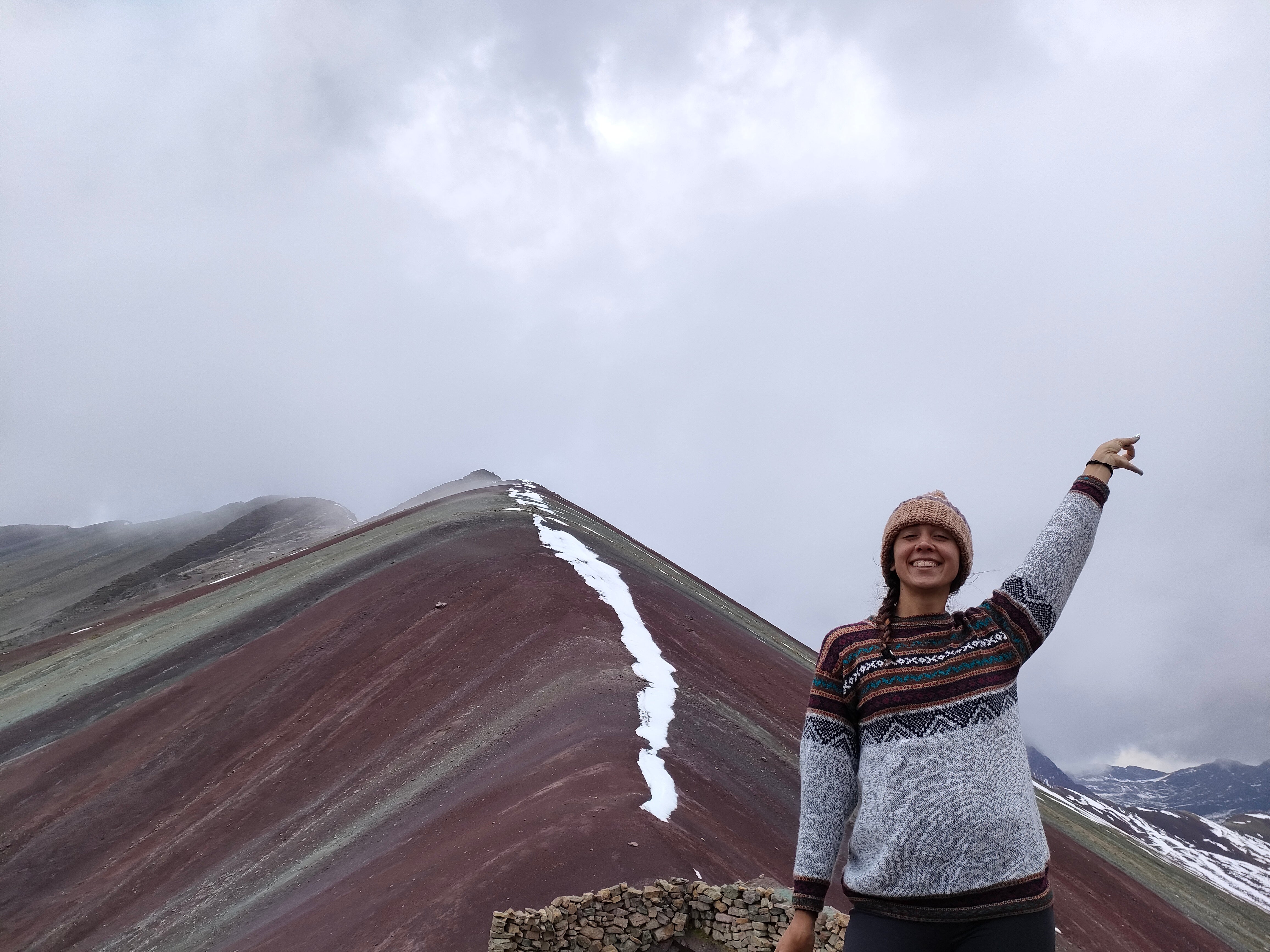 A cheerful person in a knitted beanie and sweater standing in front of the Rainbow Mountain in Peru, playfully pointing to the sky with the unique colorful striations of the mountain visible in the background.