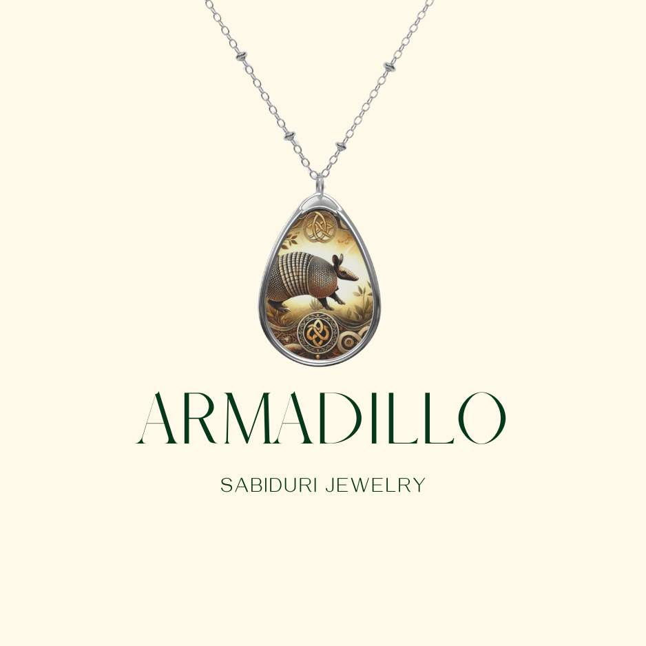 A delicate silver necklace with a teardrop-shaped pendant showcasing an armadillo surrounded by earthy-toned engravings. The pendant is set against a neutral background, with the text "ARMADILLO" and "SABIDURI JEWELRY" elegantly positioned below.