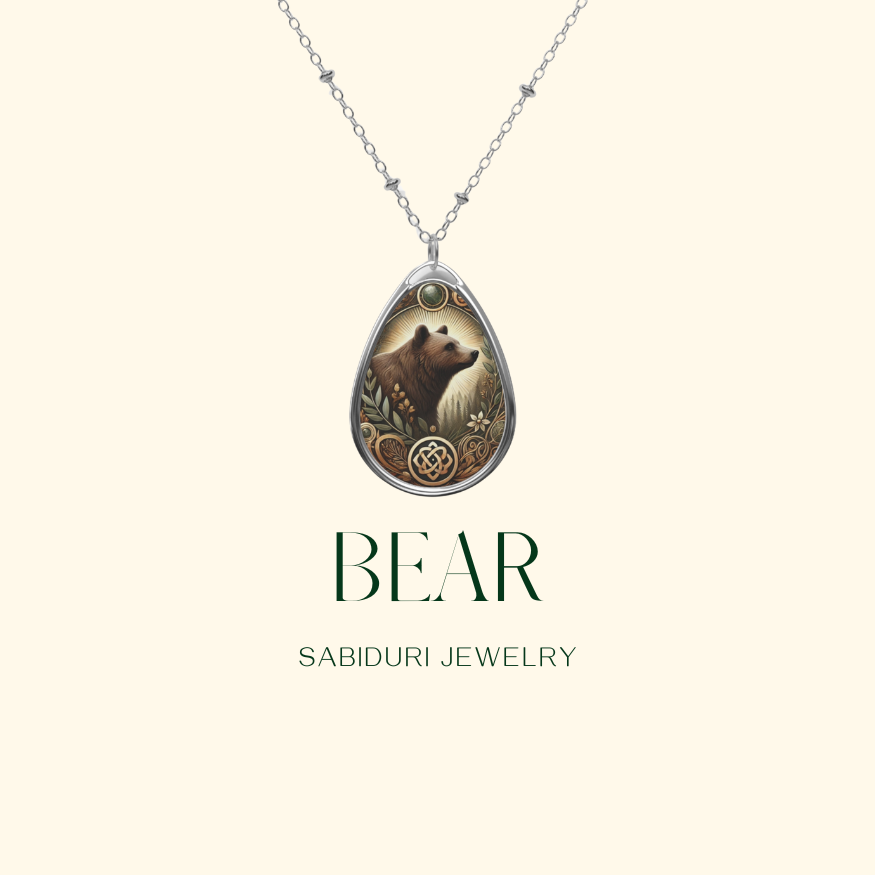 A delicate silver necklace with a teardrop-shaped pendant showcasing a bear illustration, detailed with floral and Celtic-inspired engravings. The pendant is displayed on a soft, neutral background, with the text "BEAR" and "SABIDURI JEWELRY" elegantly positioned beneath.