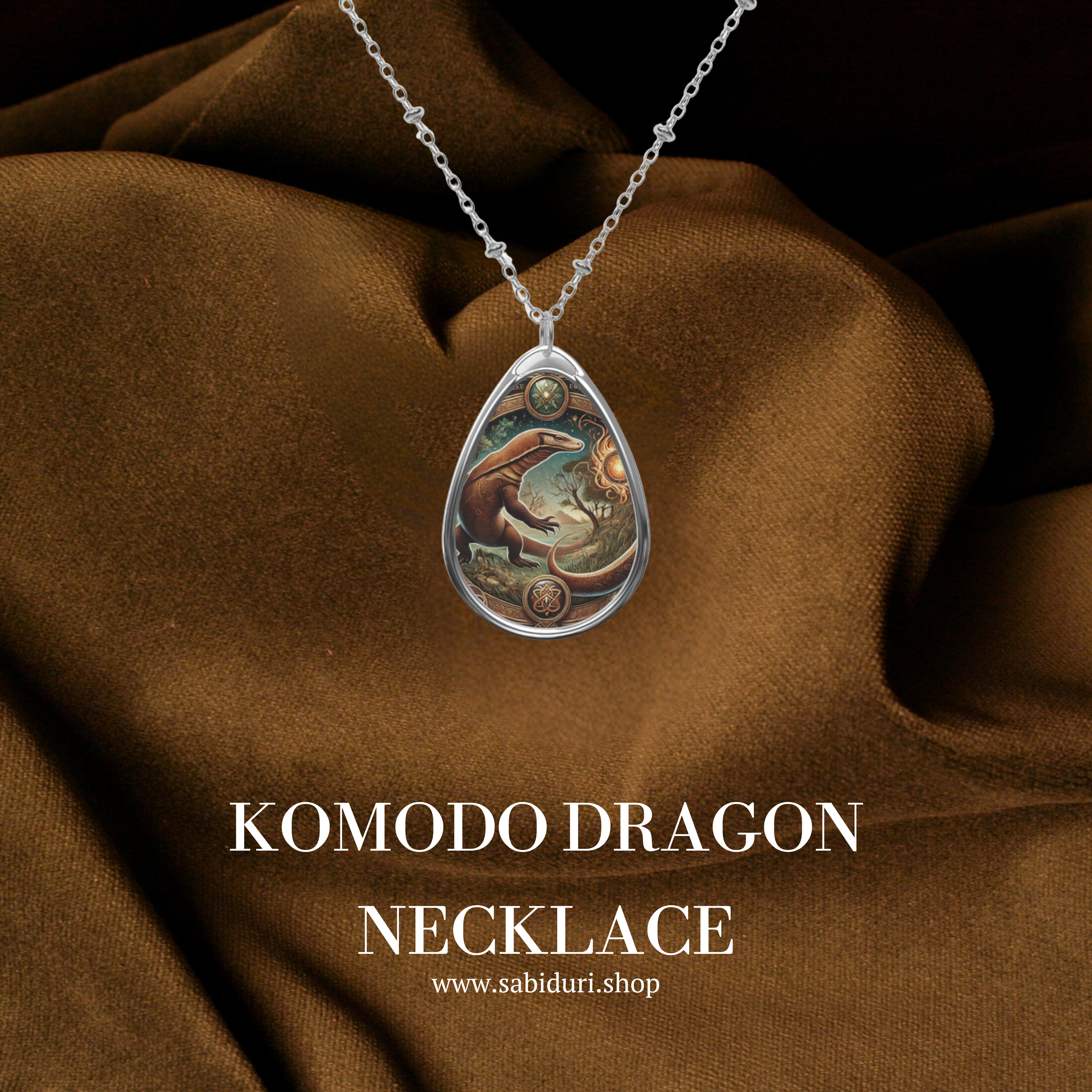 A silver teardrop-shaped pendant featuring an intricately designed Komodo dragon with earthy tones and glowing sun motifs, suspended from a delicate chain against rich brown fabric. The text "Komodo Dragon Necklace" and "www.sabiduri.shop" appear below.