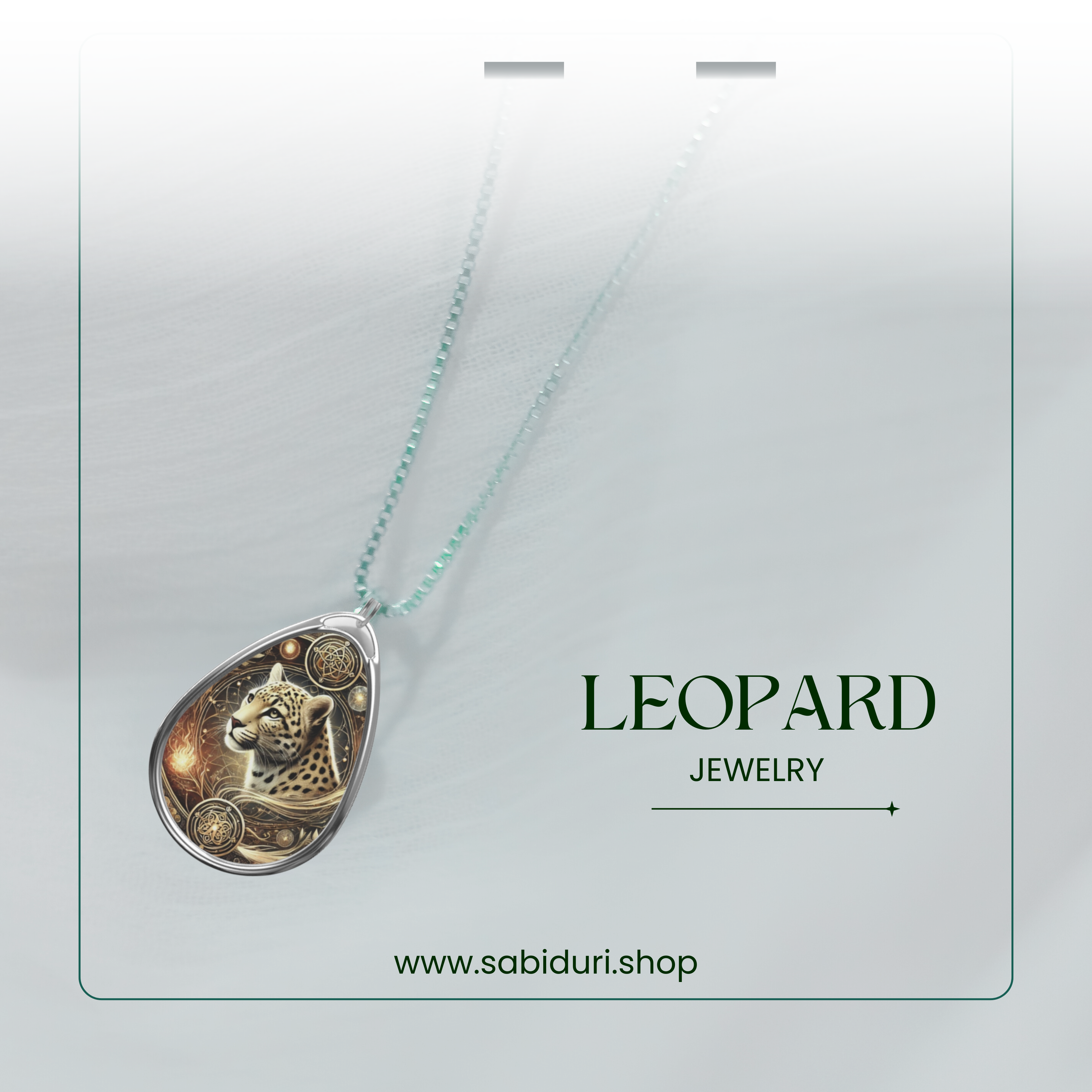 A close-up of a silver teardrop pendant adorned with a beautifully detailed leopard illustration, surrounded by intricate golden embellishments. The pendant hangs from a silver chain, elegantly displayed against a soft white fabric background. Below, "LEOPARD JEWELRY" is written in a refined font, along with "www.sabiduri.shop."