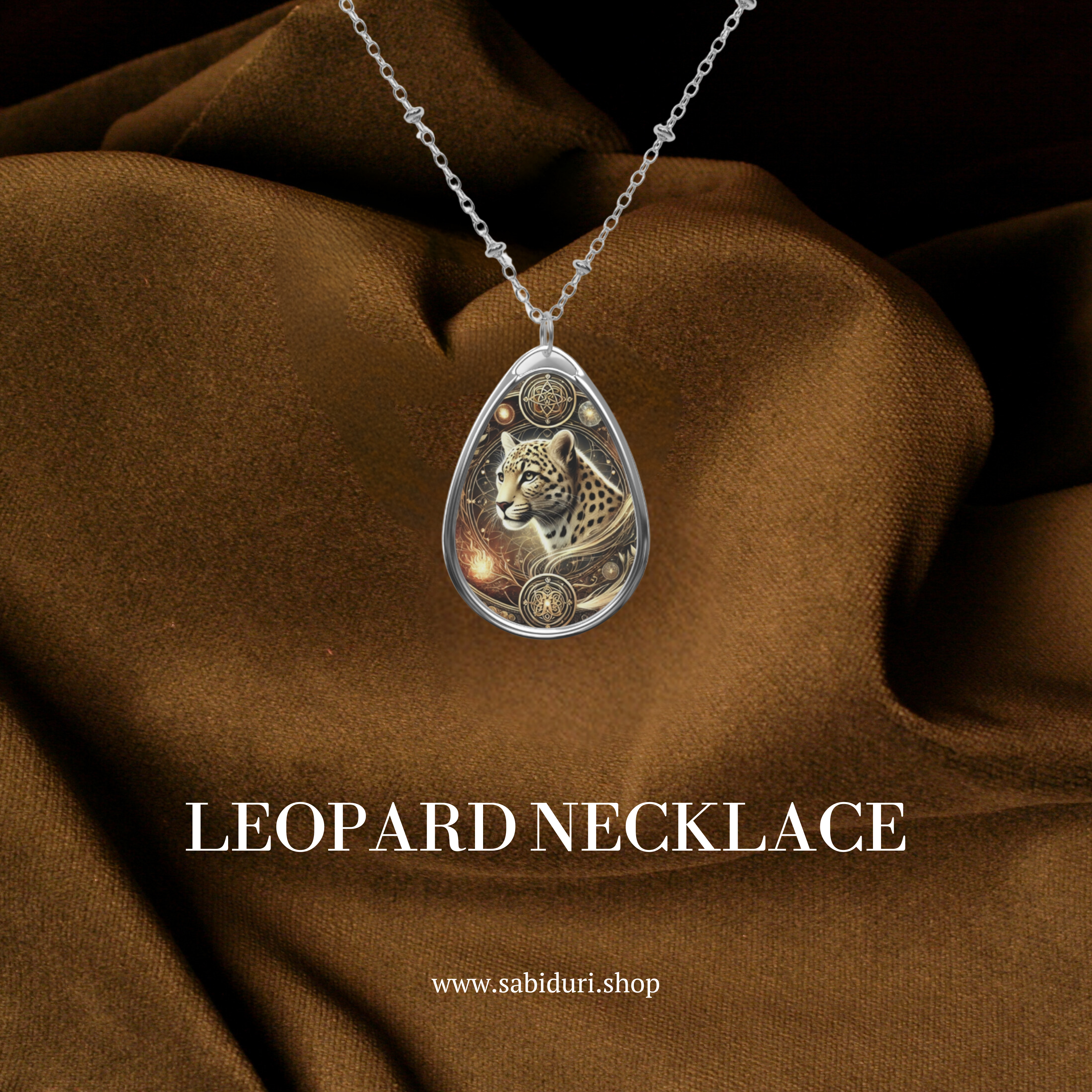 A teardrop-shaped silver pendant featuring an intricately designed leopard surrounded by celestial and ornamental patterns. The pendant hangs from a delicate silver chain, displayed against a rich brown fabric background. The text "LEOPARD NECKLACE" is prominently displayed beneath, along with the website "www.sabiduri.shop."