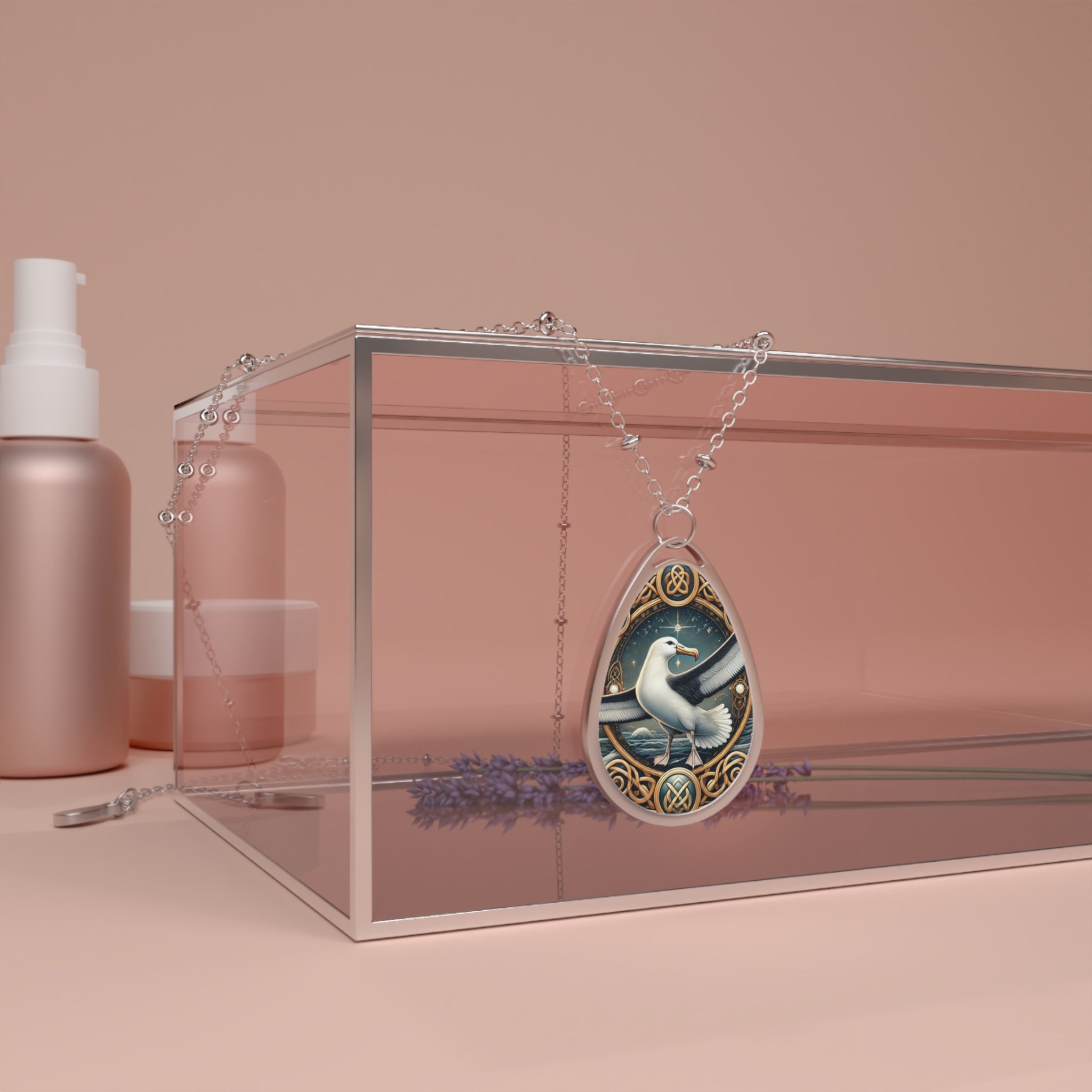 A silver chain necklace with a teardrop-shaped pendant featuring an intricately illustrated albatross soaring over ocean waves. The pendant is elegantly draped over a transparent jewelry box, surrounded by soft pink beauty product containers and sprigs of lavender in the background.