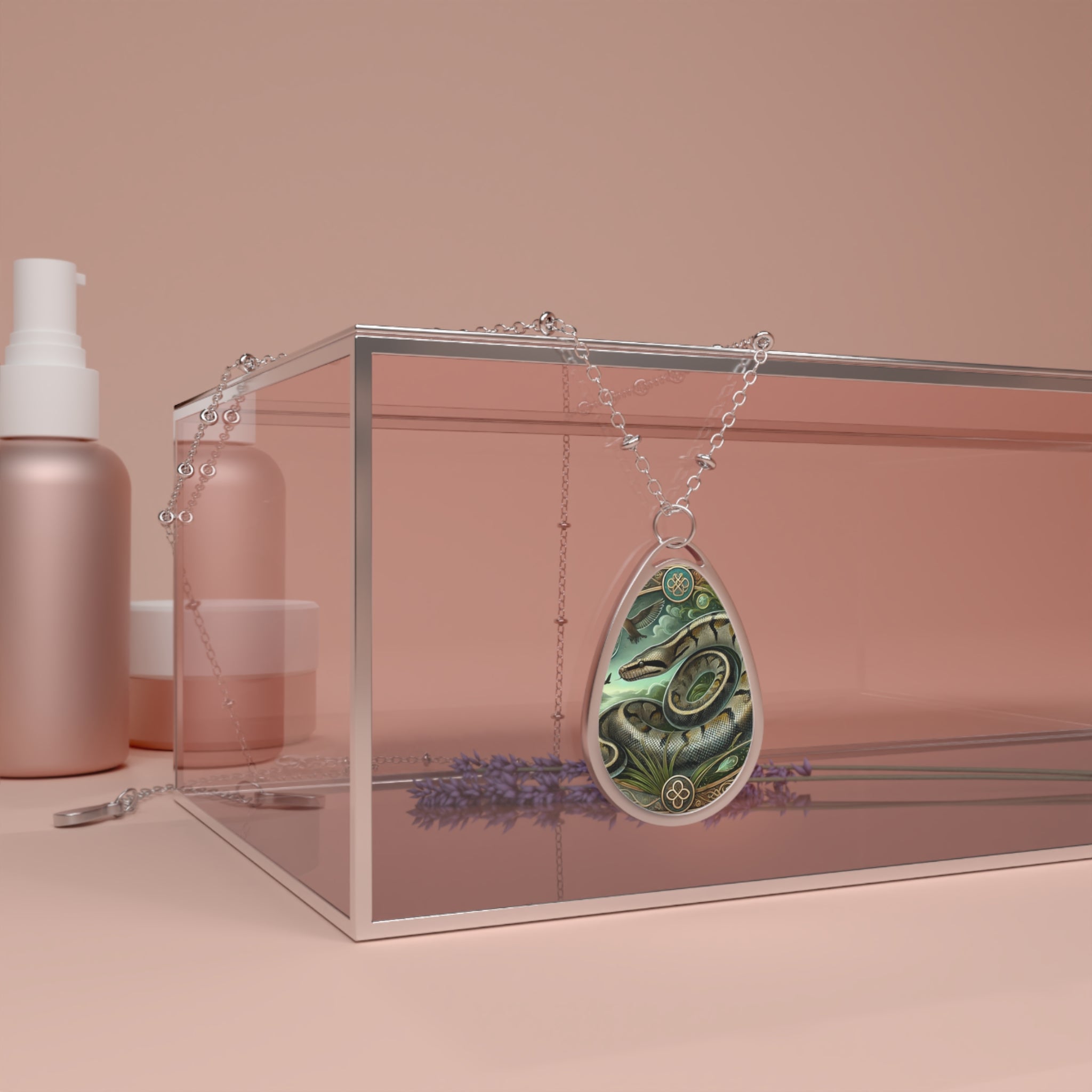 A silver chain necklace with a teardrop-shaped pendant featuring an intricately illustrated anaconda coiled in a lush jungle setting. The pendant is elegantly draped over a transparent jewelry box, surrounded by soft pink beauty product containers and sprigs of lavender in the background.