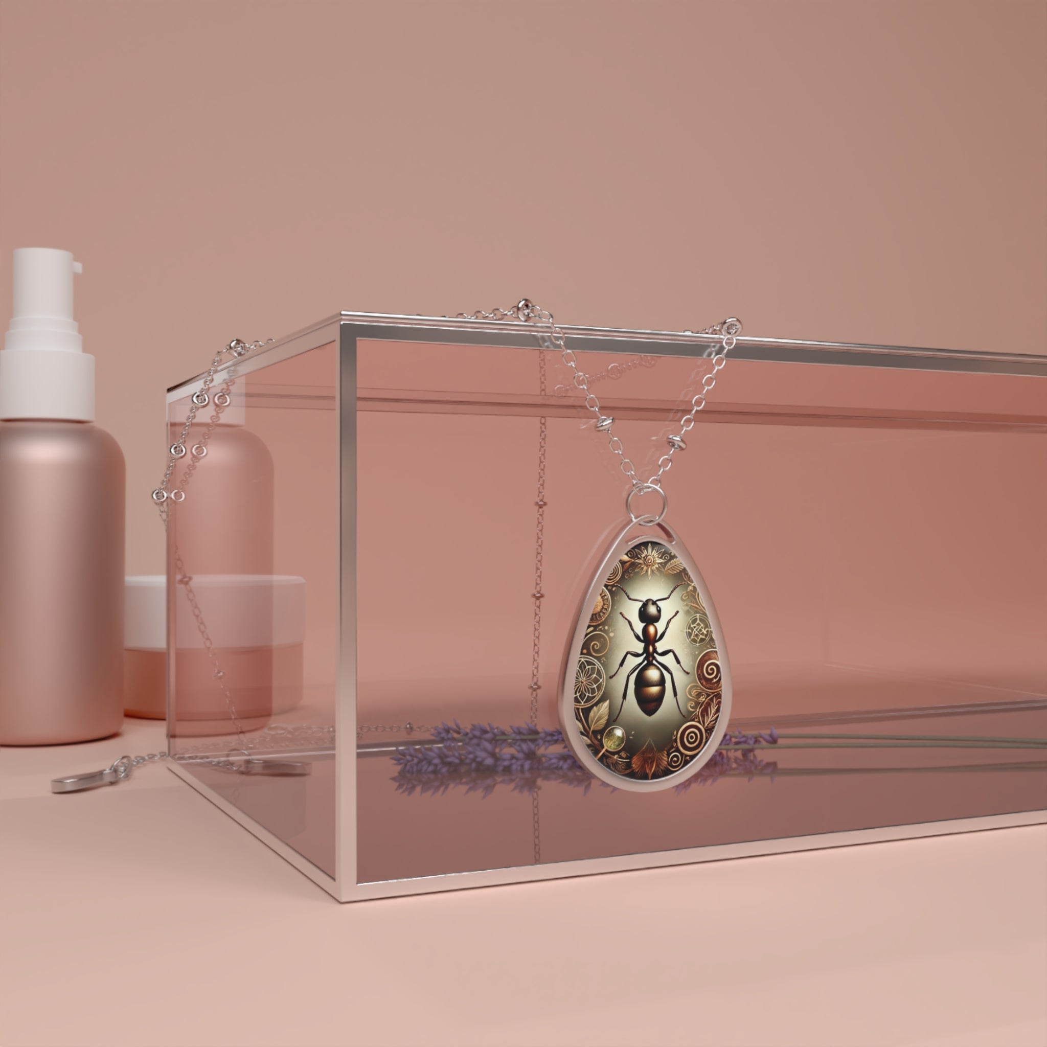 A silver chain necklace with a teardrop-shaped pendant featuring an intricately detailed ant illustration. The pendant is elegantly draped over a transparent jewelry box, surrounded by soft pink beauty product containers and sprigs of lavender in the background.