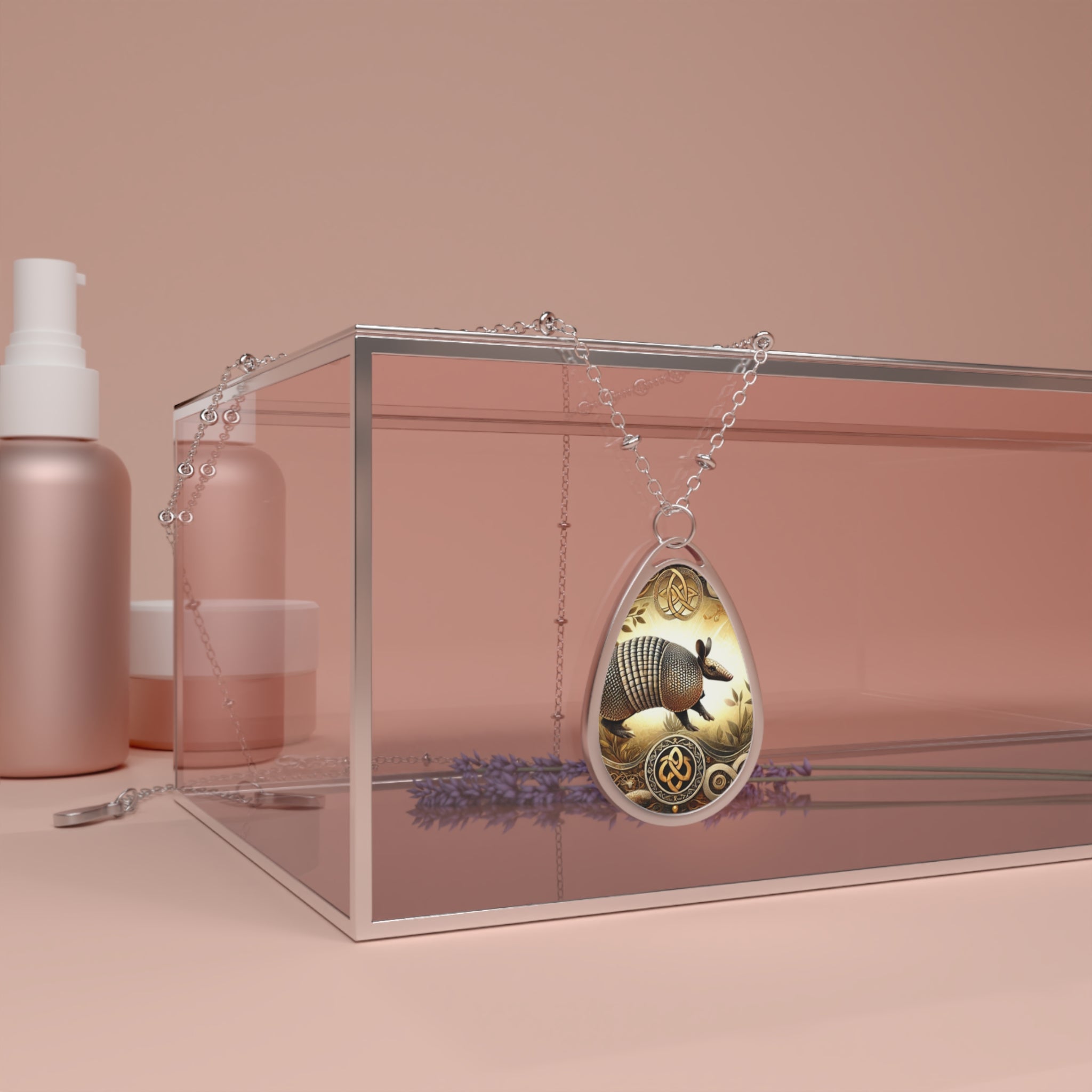 A silver chain necklace with a teardrop-shaped pendant featuring an intricately engraved armadillo design in gold and yellow tones. The pendant is elegantly draped over a modern transparent jewelry box, with soft pink beauty product containers and sprigs of lavender in the background.