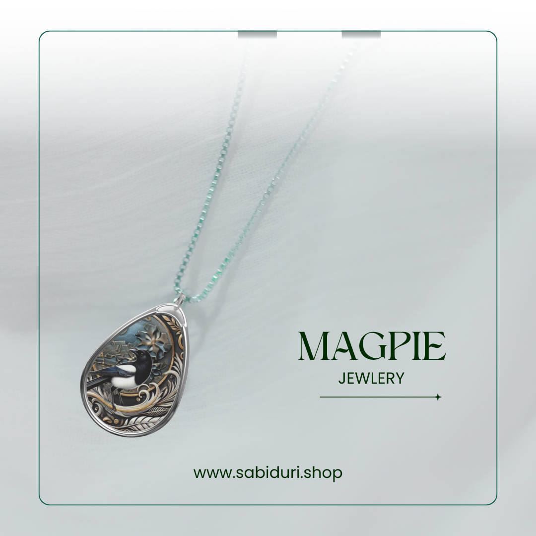 A close-up image of a teardrop-shaped pendant featuring a detailed magpie design with intricate gold and silver accents. The necklace is displayed against a soft, white textured background. The text "MAGPIE JEWELRY" is prominently shown in green, with the URL "www.sabiduri.shop" at the bottom. The image has a minimalistic and elegant presentation, framed by a thin green border.