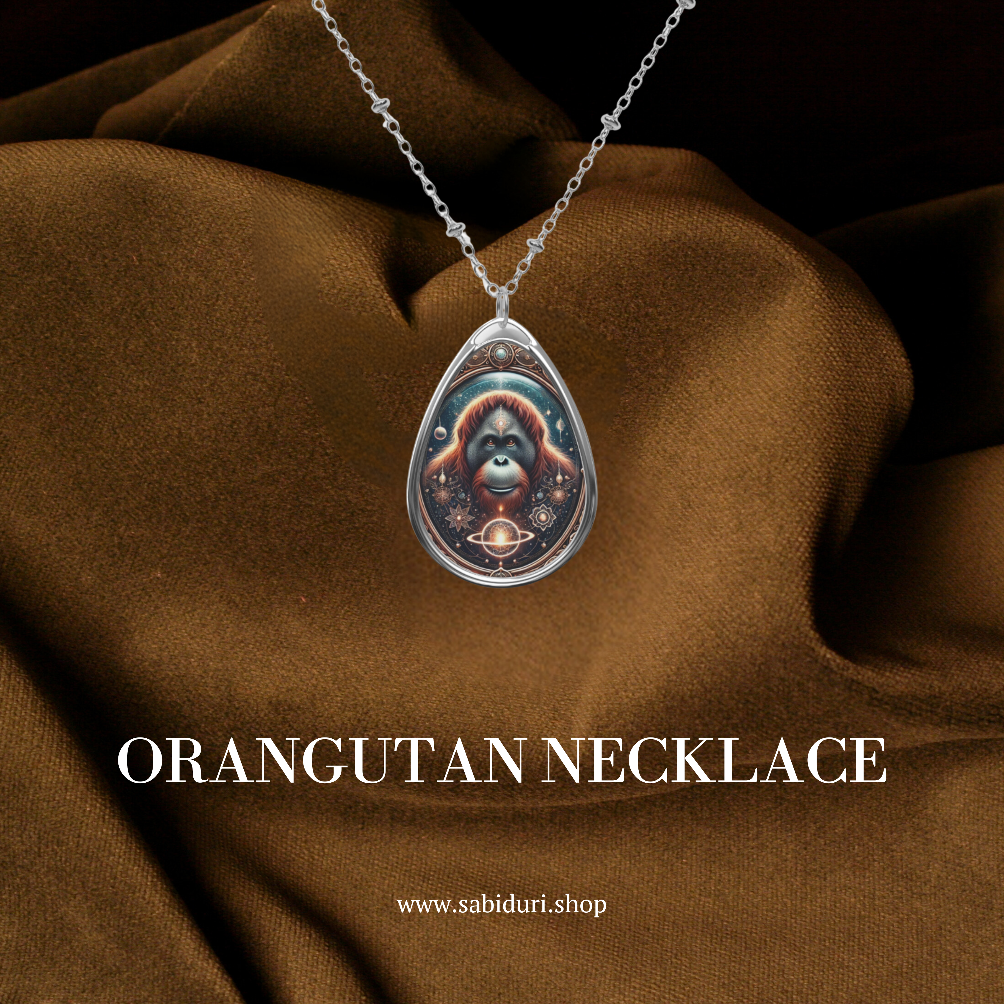 A teardrop-shaped pendant necklace featuring a detailed illustration of an orangutan with a celestial-themed background, including planets and intricate golden patterns. The necklace hangs against a rich brown fabric backdrop, emphasizing its intricate details. The text "ORANGUTAN NECKLACE" and "www.sabiduri.shop" appear below.