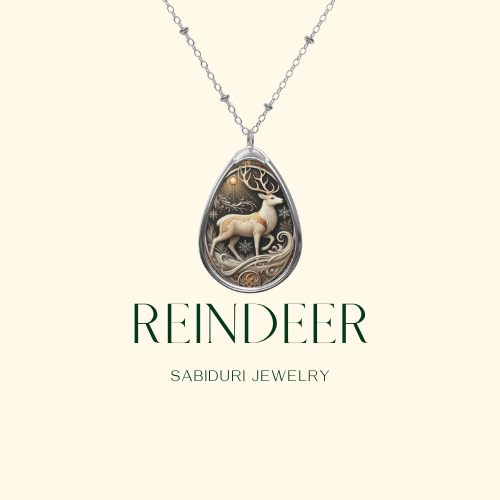 A reindeer pendant necklace displayed on a minimalist cream-colored background. The pendant highlights a beautifully designed reindeer with ornate antlers, surrounded by delicate patterns. The text reads "REINDEER Sabiduri Jewelry.