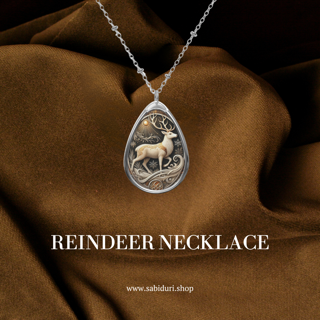 A close-up of a reindeer pendant necklace on rich brown fabric. The pendant features a gracefully posed reindeer with intricate detailing in the antlers and wintry accents, evoking a sense of wilderness and winter. The text reads "REINDEER Necklace" along with the website "www.sabiduri.shop.