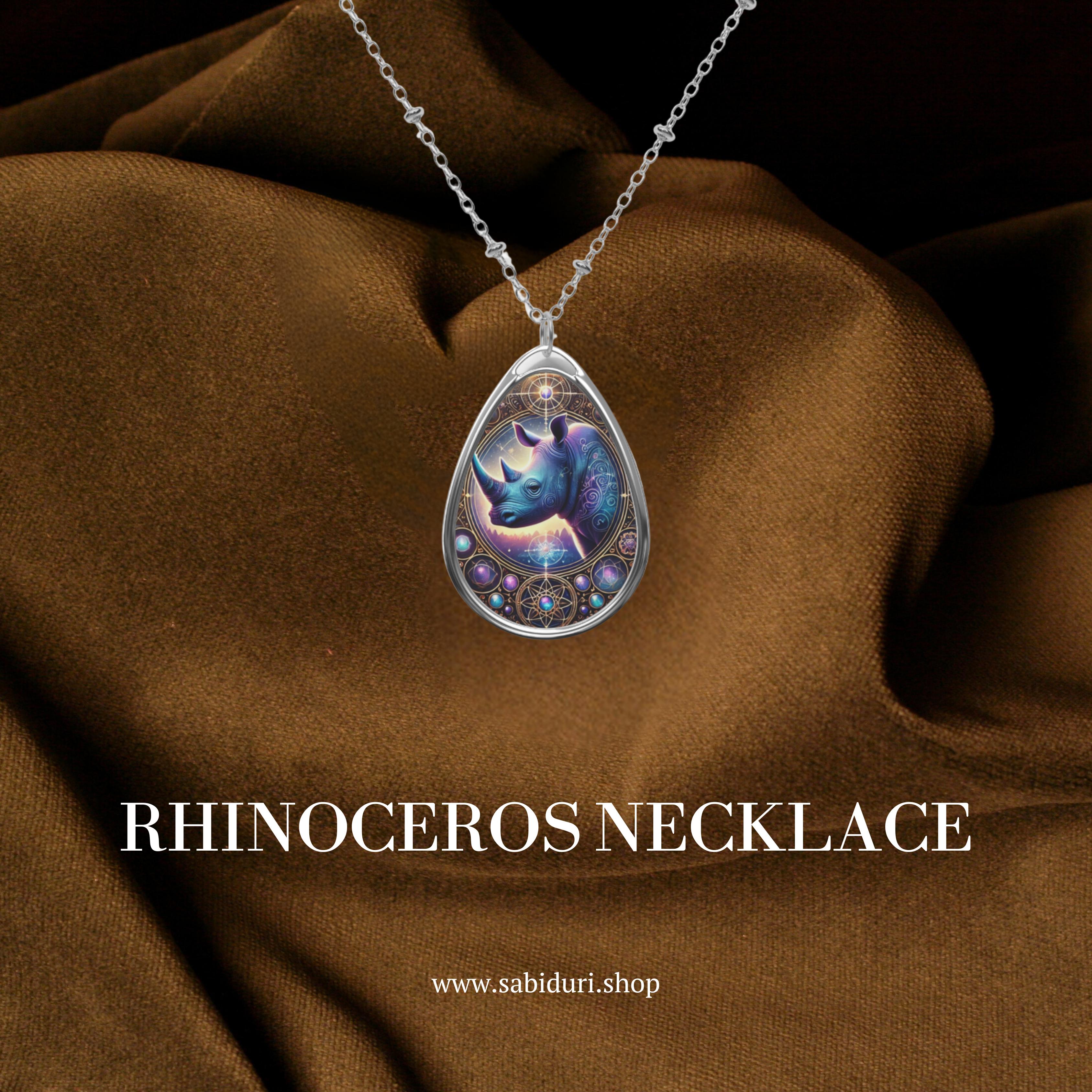 A silver teardrop-shaped pendant necklace featuring a beautifully detailed rhinoceros illustration with mystical purple and blue hues, adorned with intricate geometric designs. The necklace rests on a luxurious brown fabric background with "Rhinoceros Necklace" and "www.sabiduri.shop" text.