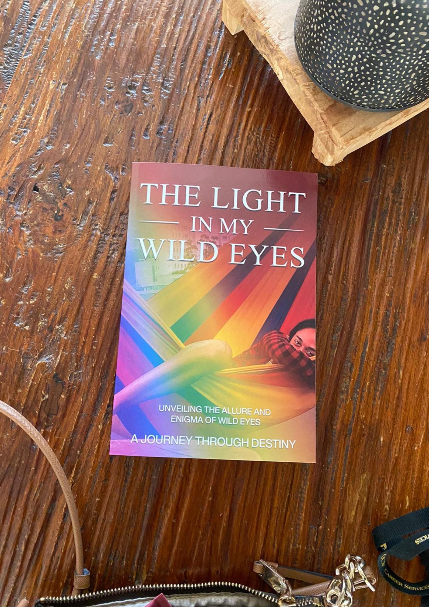 The book 'The Light in My Wild Eyes' is placed on a textured wooden surface next to a zipped pouch, with a decorative object in the background. The cover features vibrant rainbow hues with an image of a person peeking through a multicolored lens.