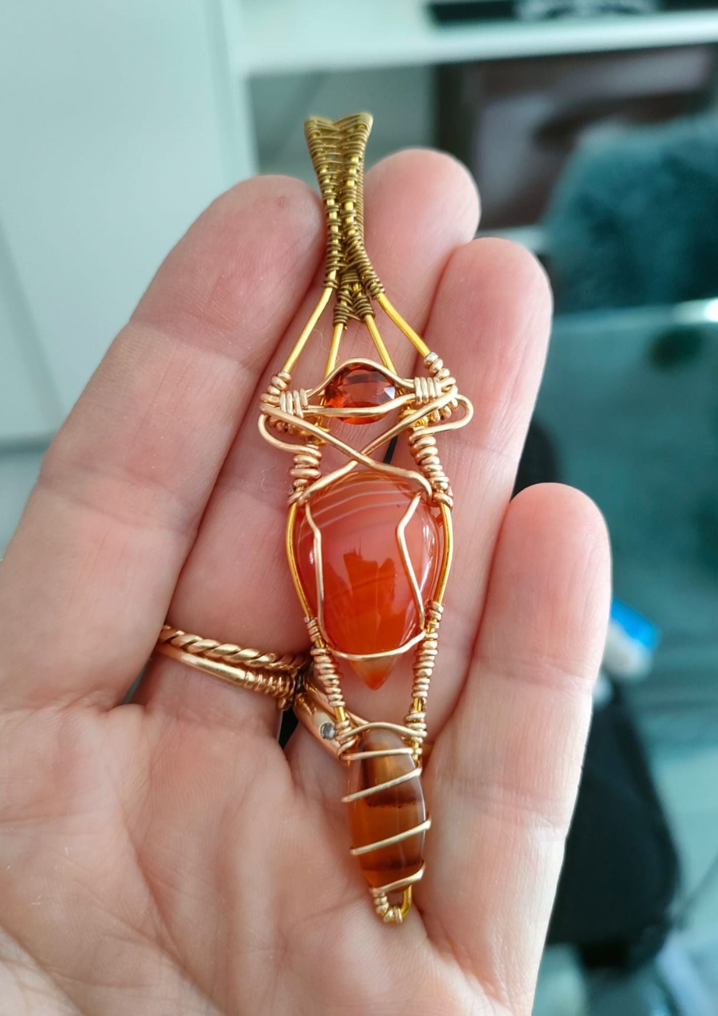 Close-up of a hand holding an intricately wire-wrapped pendant with a bright orange stone at its center, showcased against an out-of-focus indoor background.