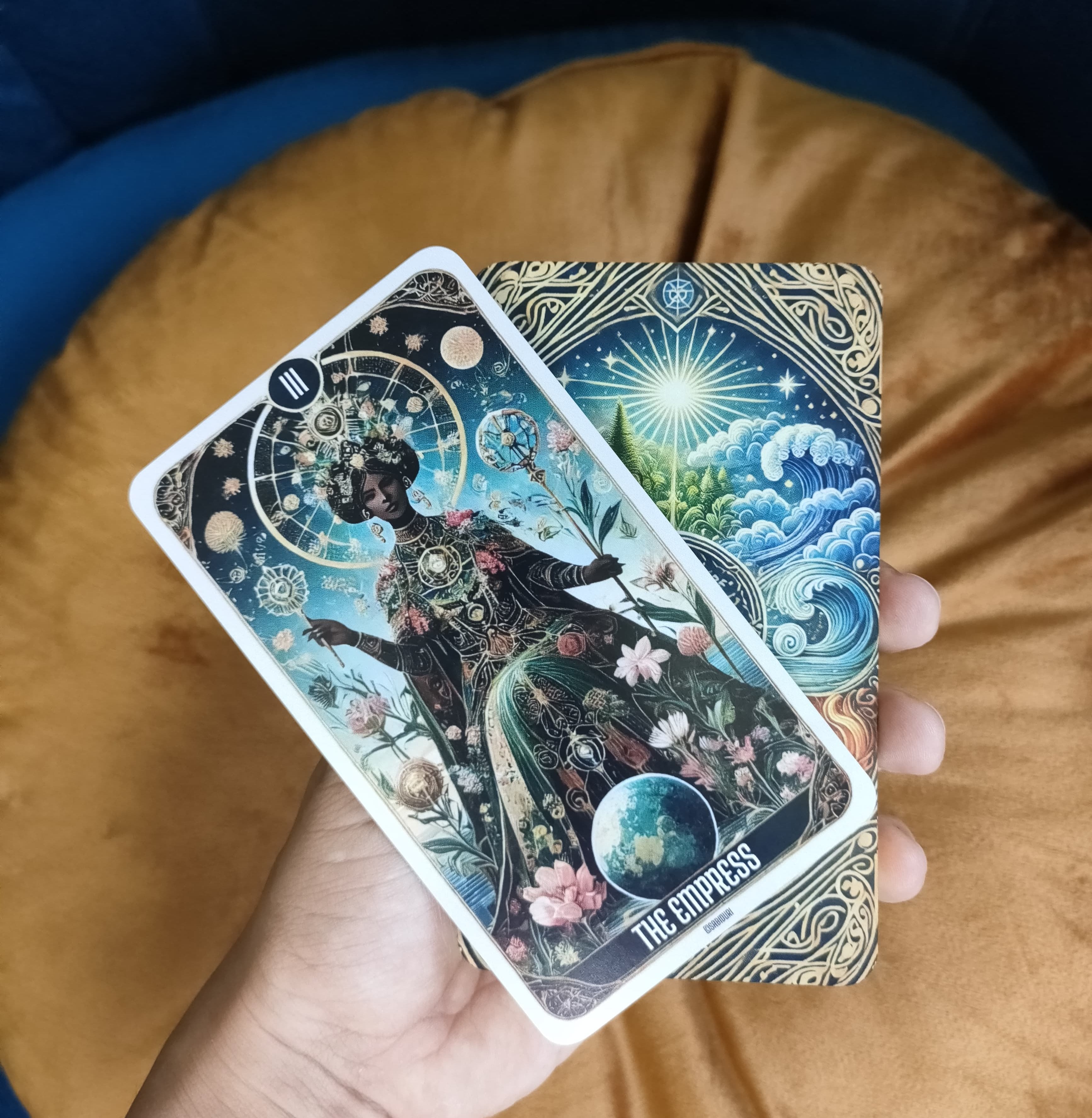 A close-up of "The Empress" tarot card from the Sabiduri deck, held in a hand. The card depicts a woman adorned with floral and celestial imagery, symbolizing fertility, abundance, and creation.