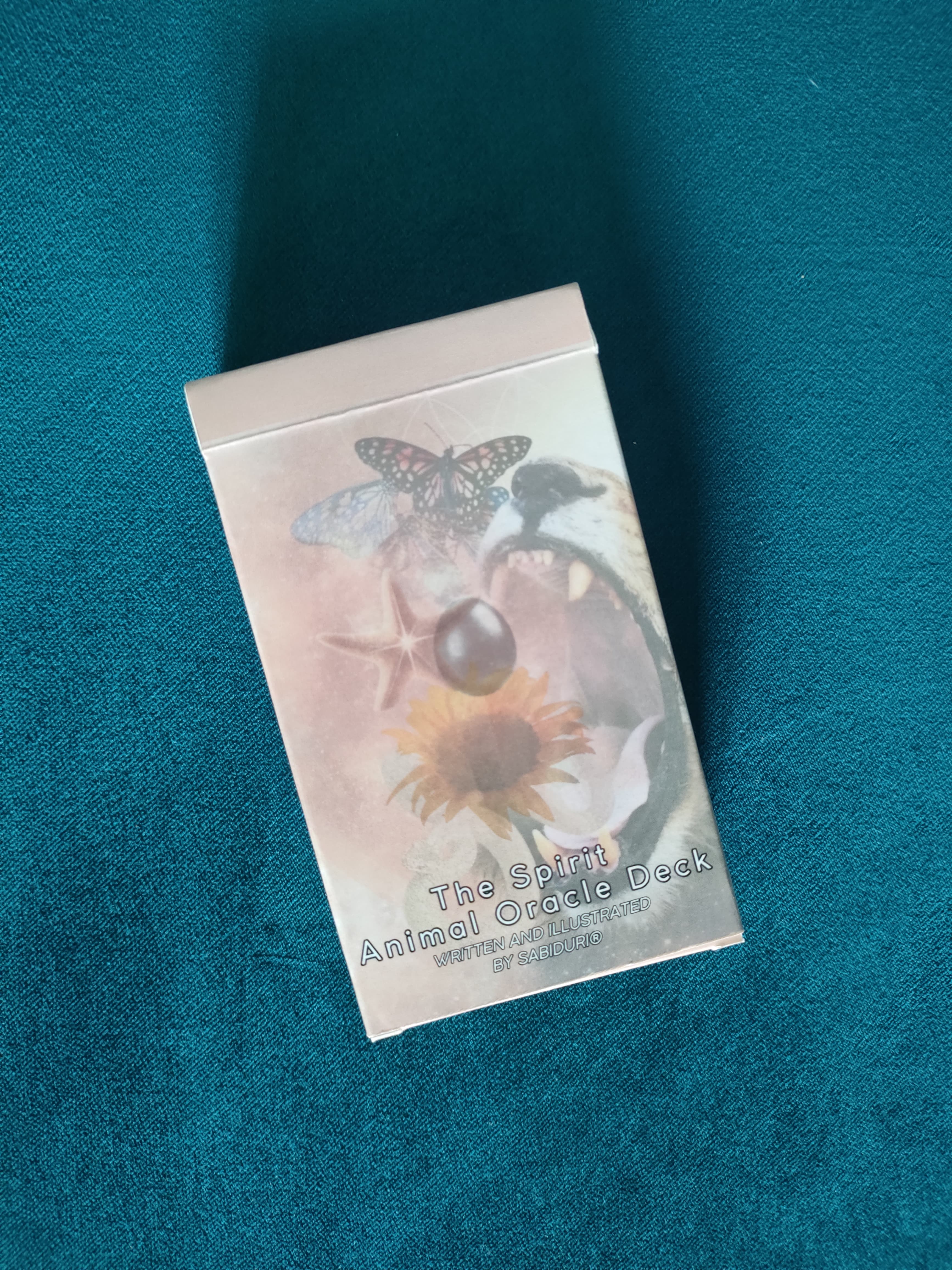 The front of The Spirit Animal Oracle Deck box, displaying a mystical illustration of a wolf, butterfly, starfish, and sunflower with a cosmic, ethereal background. The text reads: "The Spirit Animal Oracle Deck – Written and Illustrated by SABIDURI."