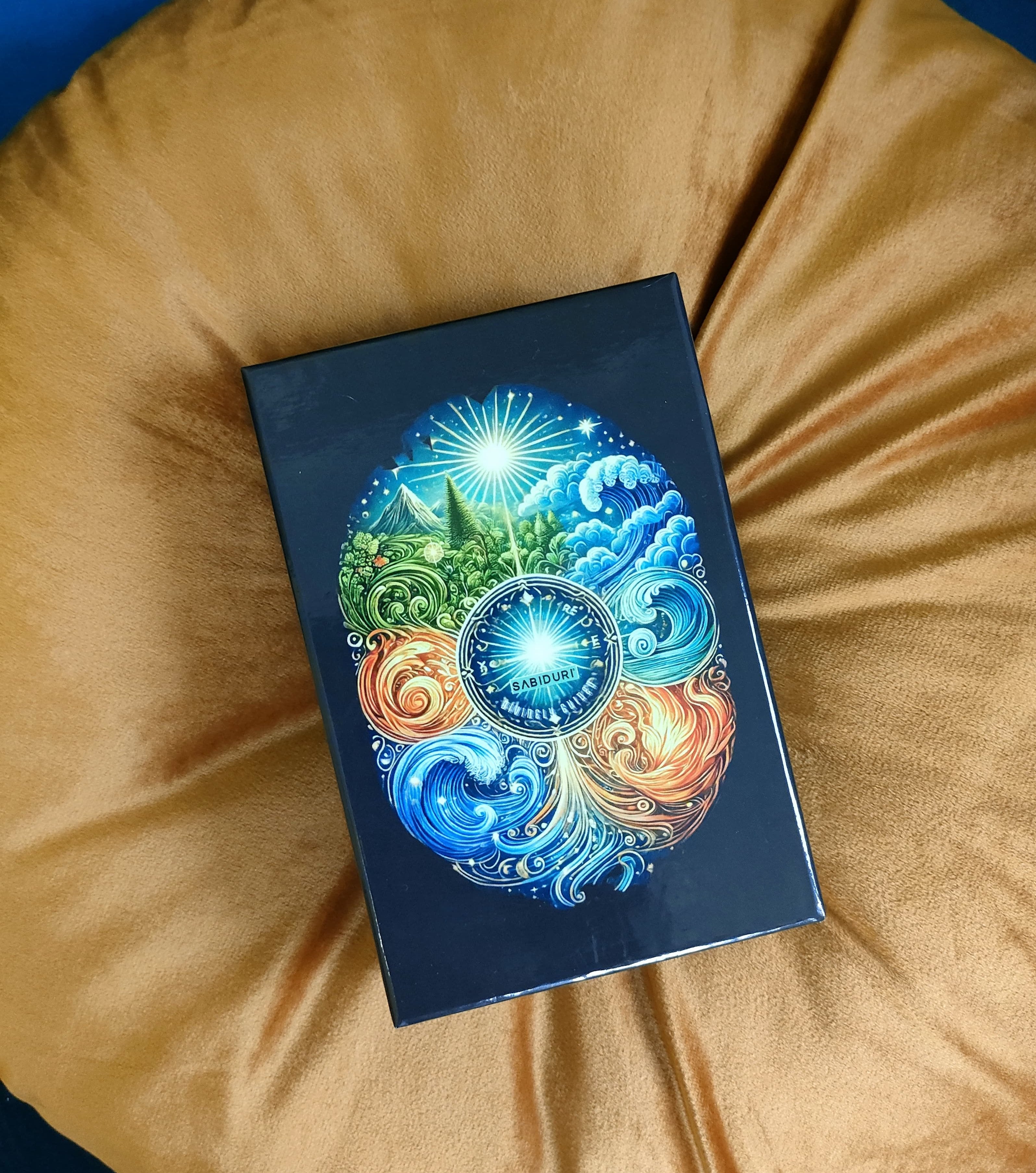 A Sabiduri Tarot deck box resting on a plush mustard-colored pillow. The box features an intricate, colorful design with swirling elements representing fire, water, air, and earth around a central circular logo.