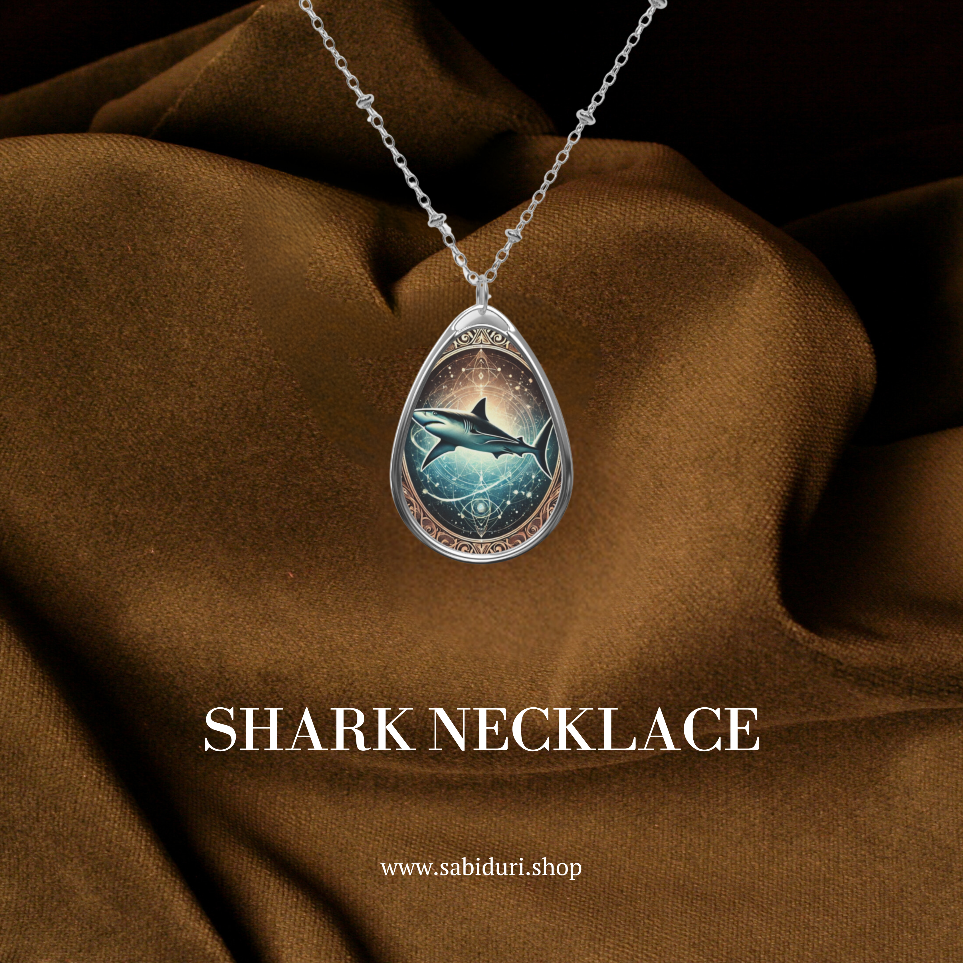 A close-up of a teardrop-shaped shark necklace pendant with an intricate celestial design, featuring a shark swimming through a cosmic ocean. The pendant hangs from a delicate silver chain against a textured brown fabric backdrop, with "SHARK NECKLACE" and "www.sabiduri.shop" text displayed below.