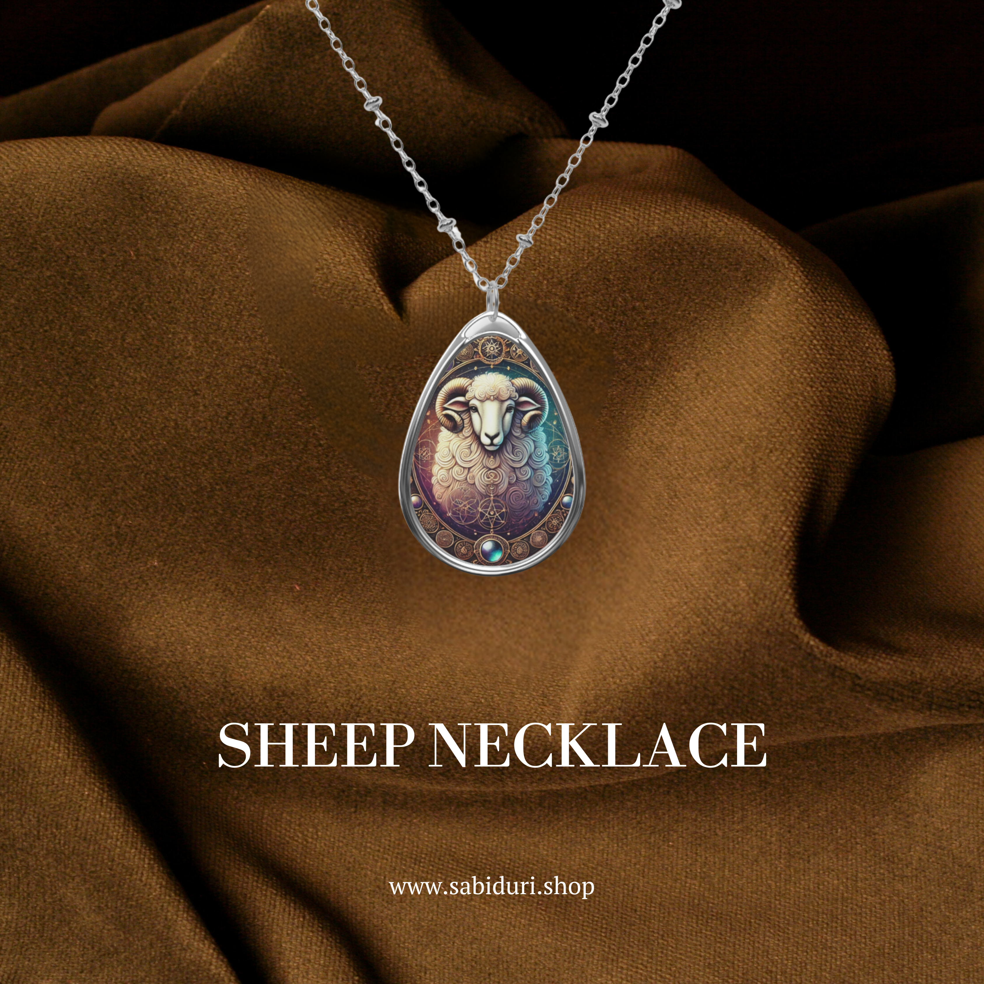 A close-up of a teardrop-shaped pendant necklace featuring an intricately designed sheep head with curled horns. The pendant has ornate golden details and a gemstone accent, set against a rich brown fabric backdrop.