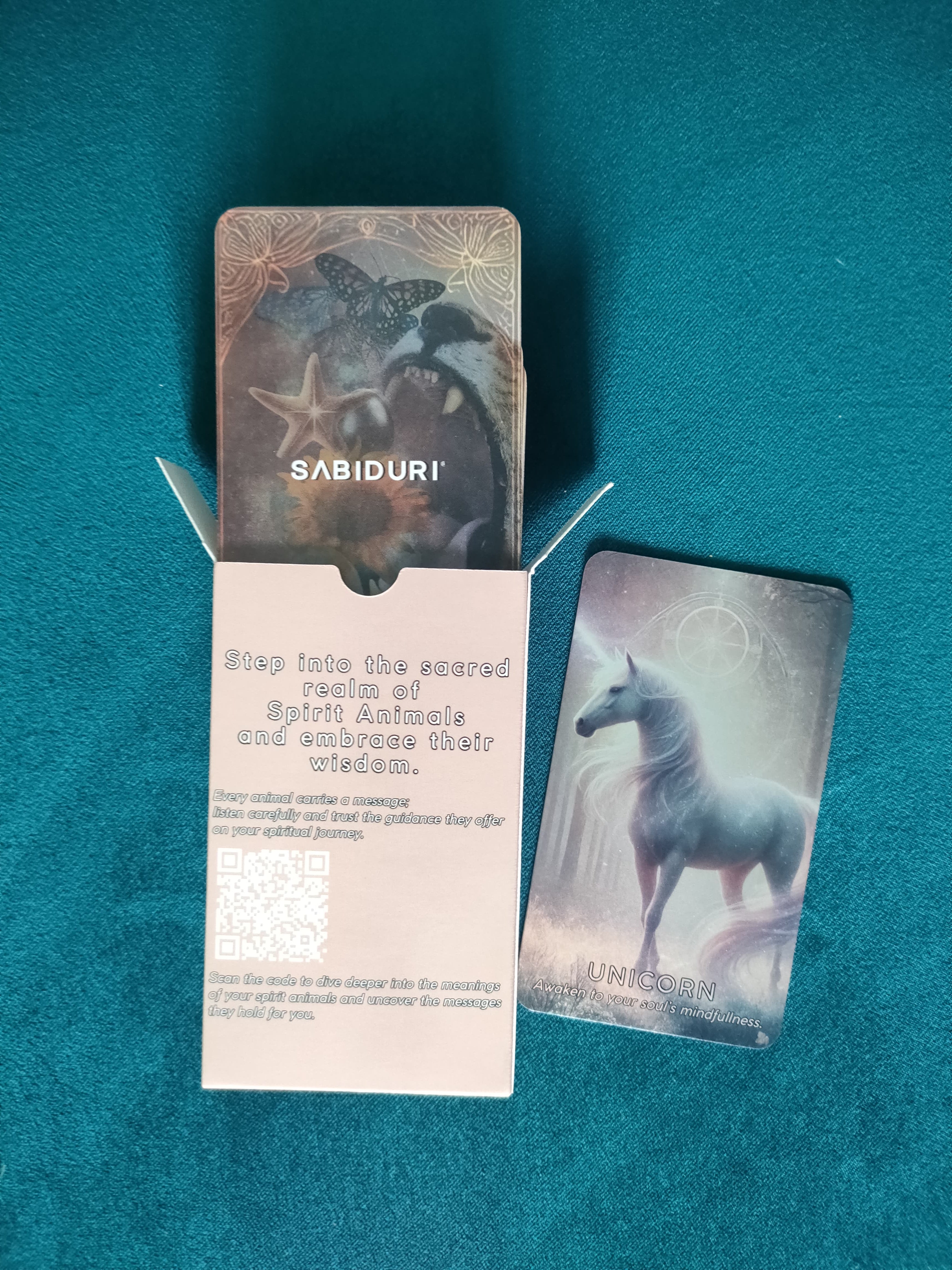 An open Spirit Animal Oracle deck box placed on a teal fabric background. A "Unicorn" card is lying next to it, displaying soft glowing artwork. Inside the box, a printed message invites users to step into the sacred realm of spirit animals, with a QR code for additional guidance.