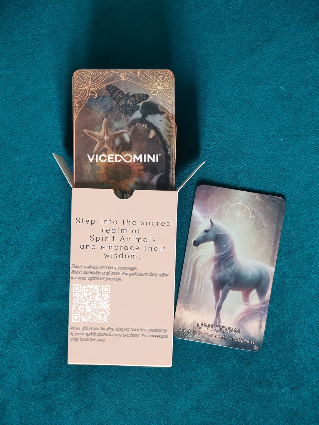 An open Spirit Animal Oracle deck box placed on a teal fabric background. A "Unicorn" card is lying next to it, displaying soft glowing artwork. Inside the box, a printed message invites users to step into the sacred realm of spirit animals, with a QR code for additional guidance.
