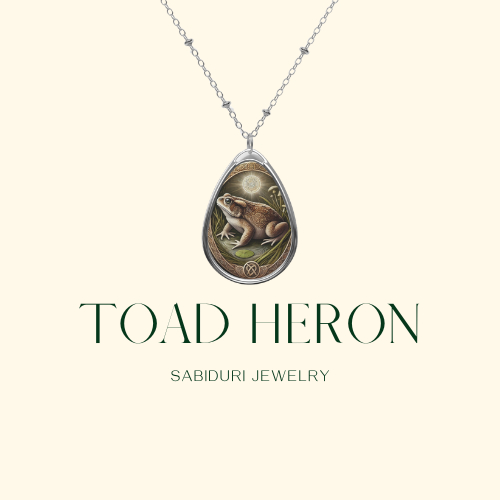 A close-up of a toad pendant necklace on a minimalist cream-colored background. The pendant features a toad surrounded by plant motifs and mystical symbols. The text reads "TOAD Sabiduri Jewelry.