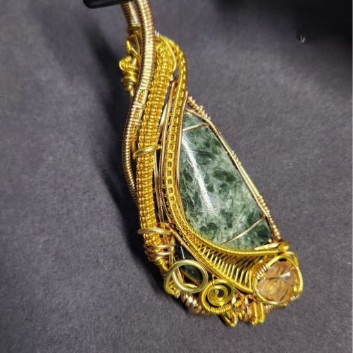 A bold, yellow-gold wire-wrapped pendant with a large green seraphinite stone accented by a small, clear quartz and a golden-hued stone on a grey background.