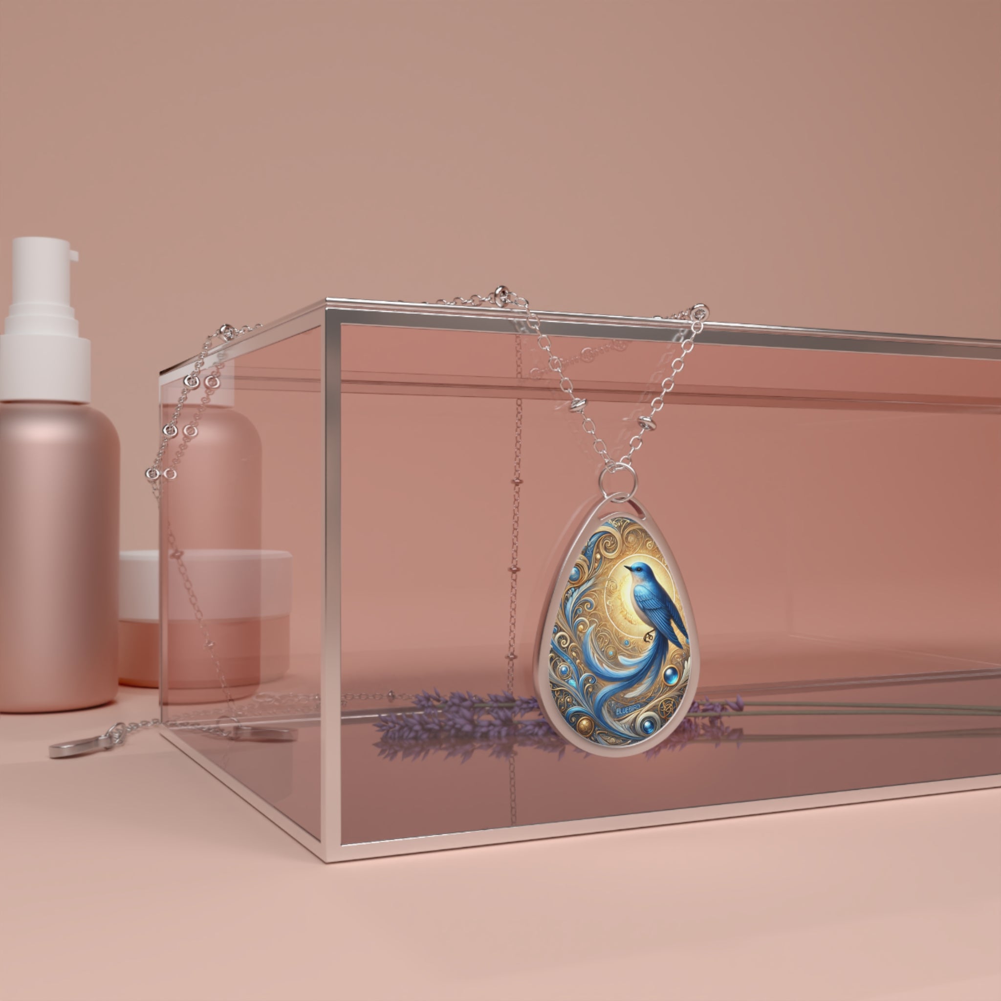 A teardrop-shaped pendant necklace featuring an intricate illustration of a bluebird perched on swirling golden filigree, glowing against a warm, golden-hued background. The pendant hangs over a reflective pink glass box, with lavender sprigs visible beneath it, creating an elegant and serene setting.