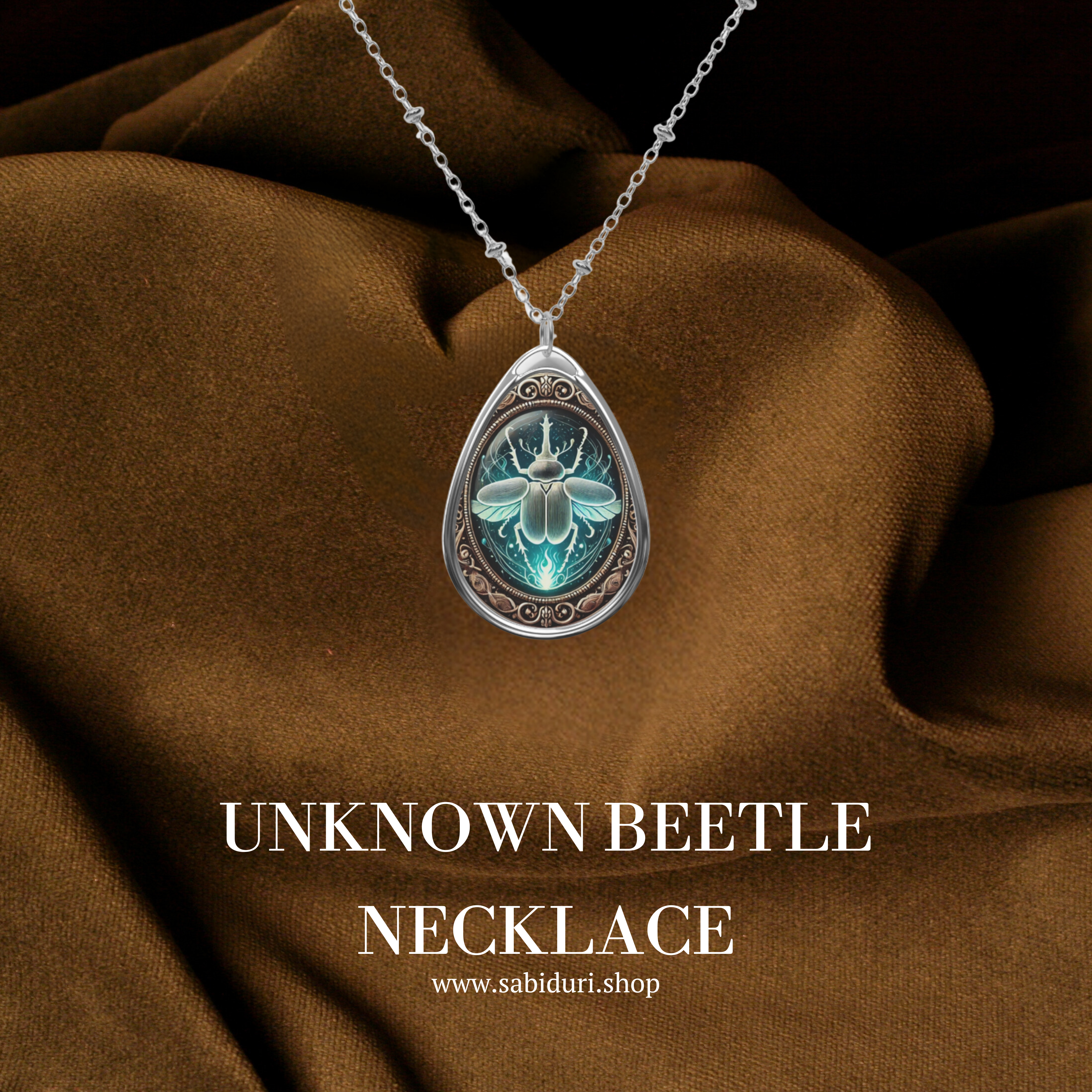 A teardrop-shaped silver necklace pendant featuring an intricate illustration of a unknown beetle surrounded by ornate golden patterns. The pendant rests on rich brown fabric, with the words "UNKNOWN BEETLE NECKLACE" and the website "www.sabiduri.shop" displayed below.