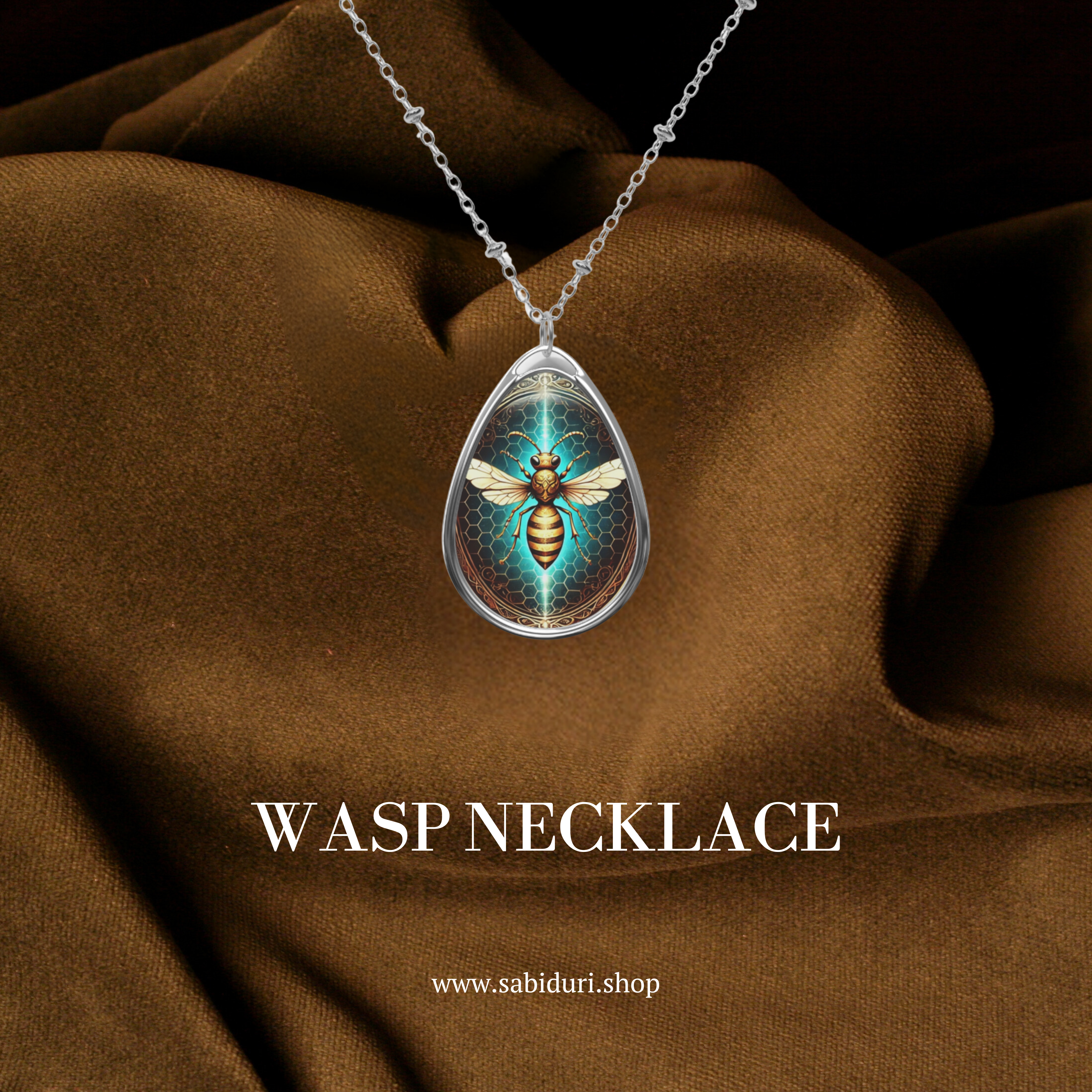 A teardrop-shaped pendant necklace featuring a detailed golden wasp with outstretched wings against a glowing honeycomb background. The pendant hangs gracefully over rich brown fabric, with the text ‘WASP NECKLACE’ and website link ‘www.sabiduri.shop’ displayed below.