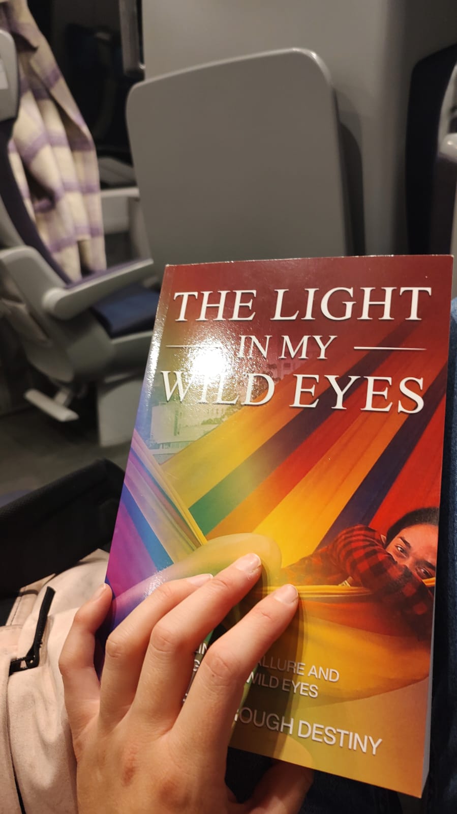 The book 'The Light in My Wild Eyes' is read during a commute, the cover's rainbow hues offering a stark contrast to the mundane surroundings, inviting introspection and adventure.