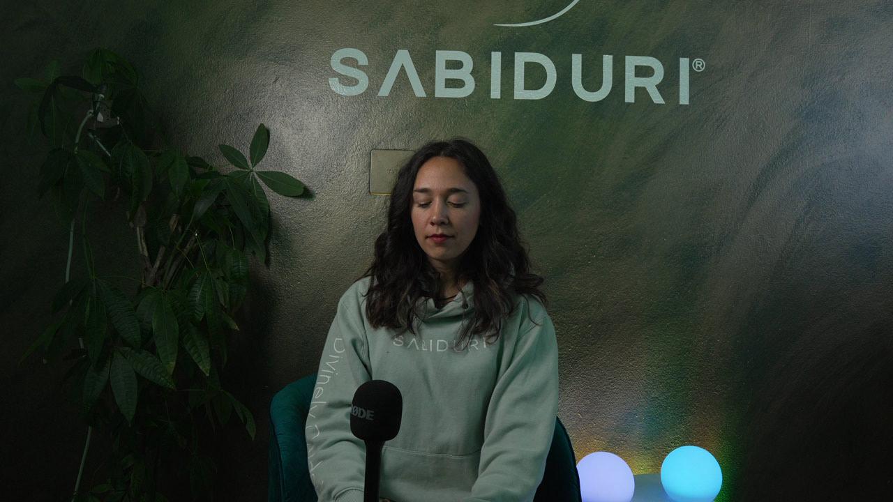 Woman sitting with a microphone in front of a green wall with 'SABIDURI' branding.