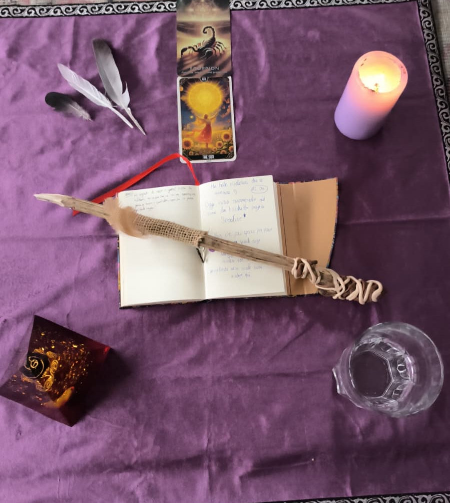 A styled flat-lay of a tarot card deck with an open book, a wooden quill pen, feathers, a lit candle, and a glass on a purple cloth.
