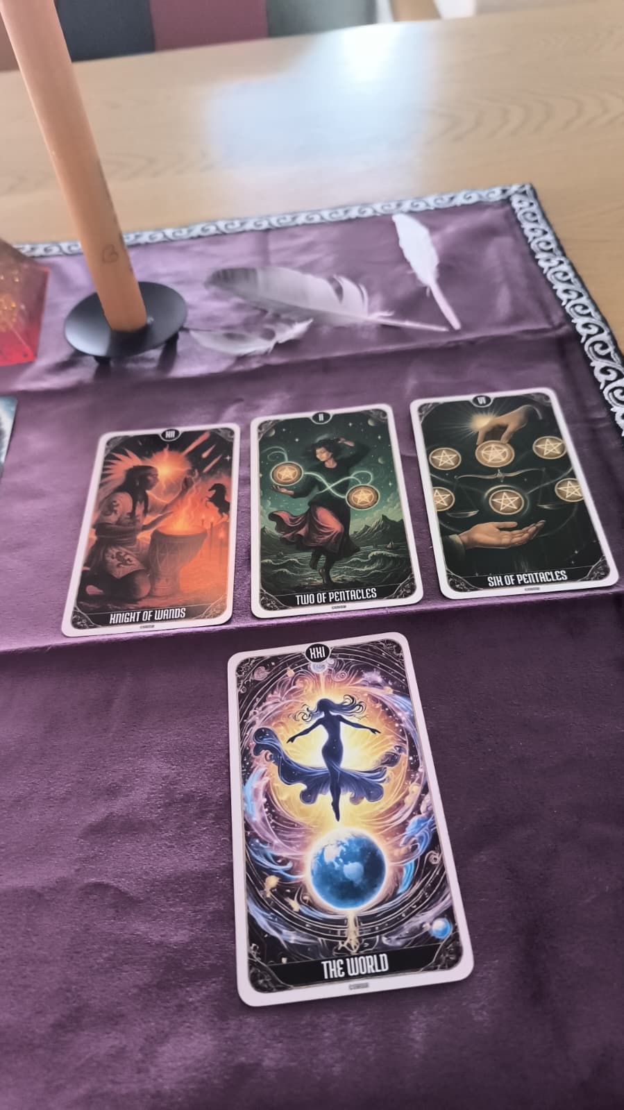 A tarot card oracle deck set up on a purple cloth on a wooden table, with four cards laid out and 'The World' card in the foreground. A candlestick and feather quill are visible in the background.