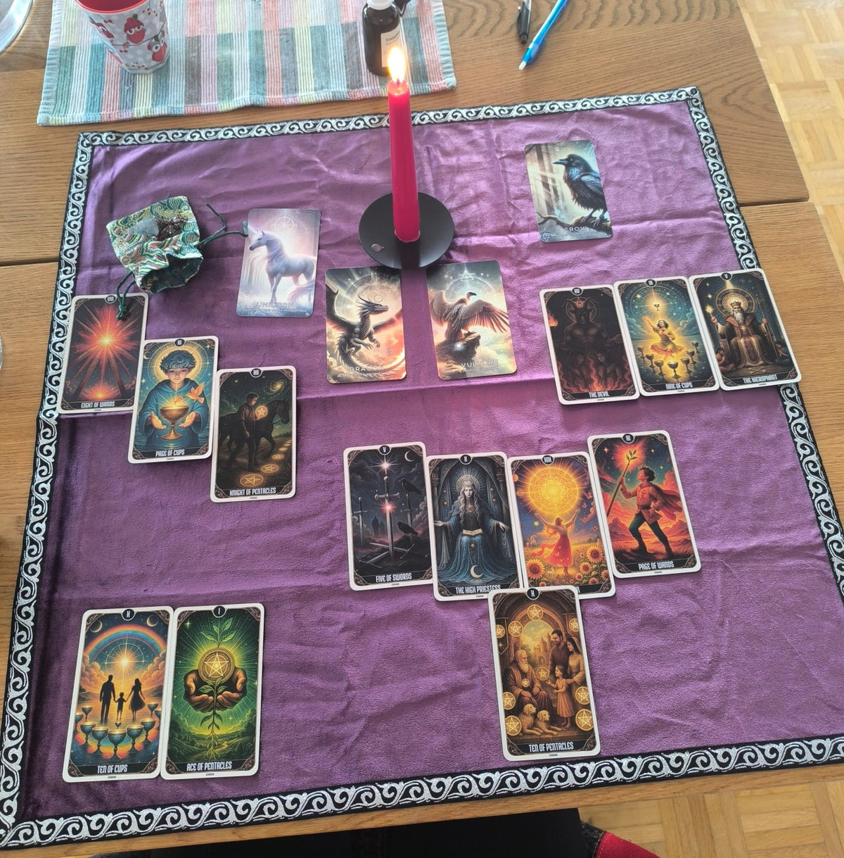 A tarot card spread arranged on a purple cloth with a decorative border, set on a wooden table with a lit red candle, a small bag, and other ritual or card elements visible.