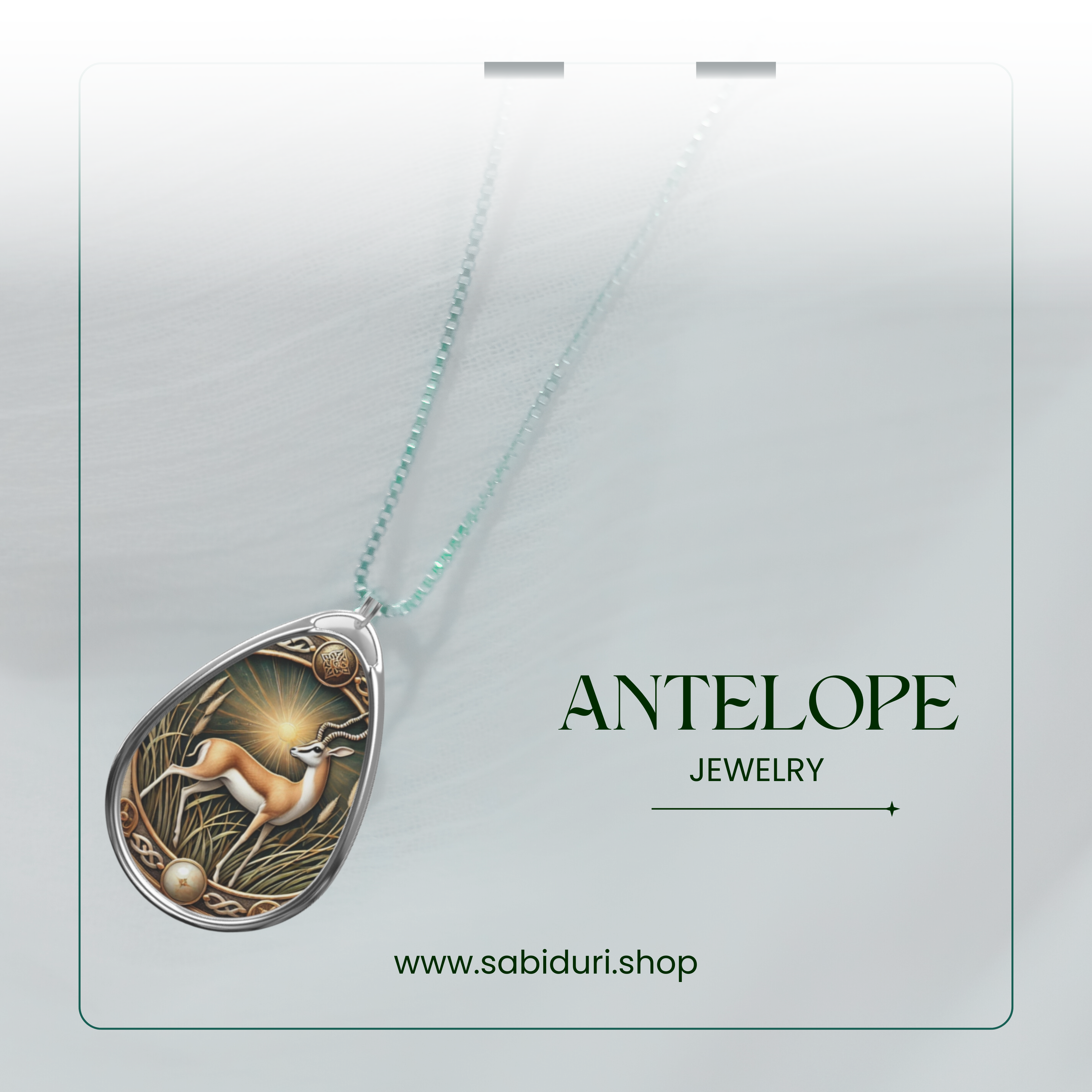 A close-up of a silver antelope pendant necklace, featuring an intricate nature-inspired design with warm golden accents. The pendant rests on a soft white fabric background, with the words "ANTELOPE JEWELRY" and the website "www.sabiduri.shop" in an elegant green font.