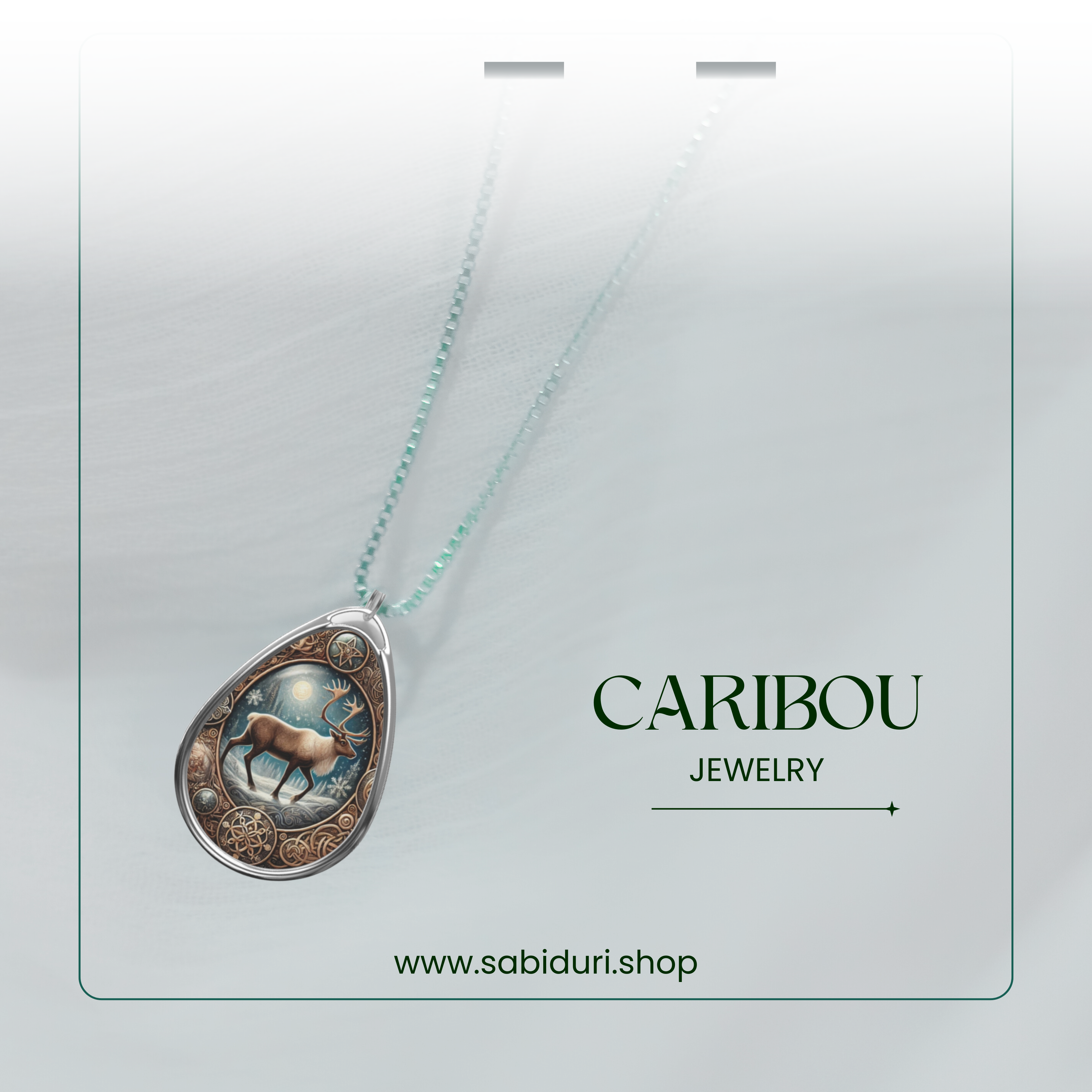 A delicate Caribou Pendant Necklace photographed against sheer white fabric. The pendant has an intricate design of a caribou under a wintery night sky, with a gold and blue color scheme. The text "CARIBOU JEWELRY" and "www.sabiduri.shop" is visible below.