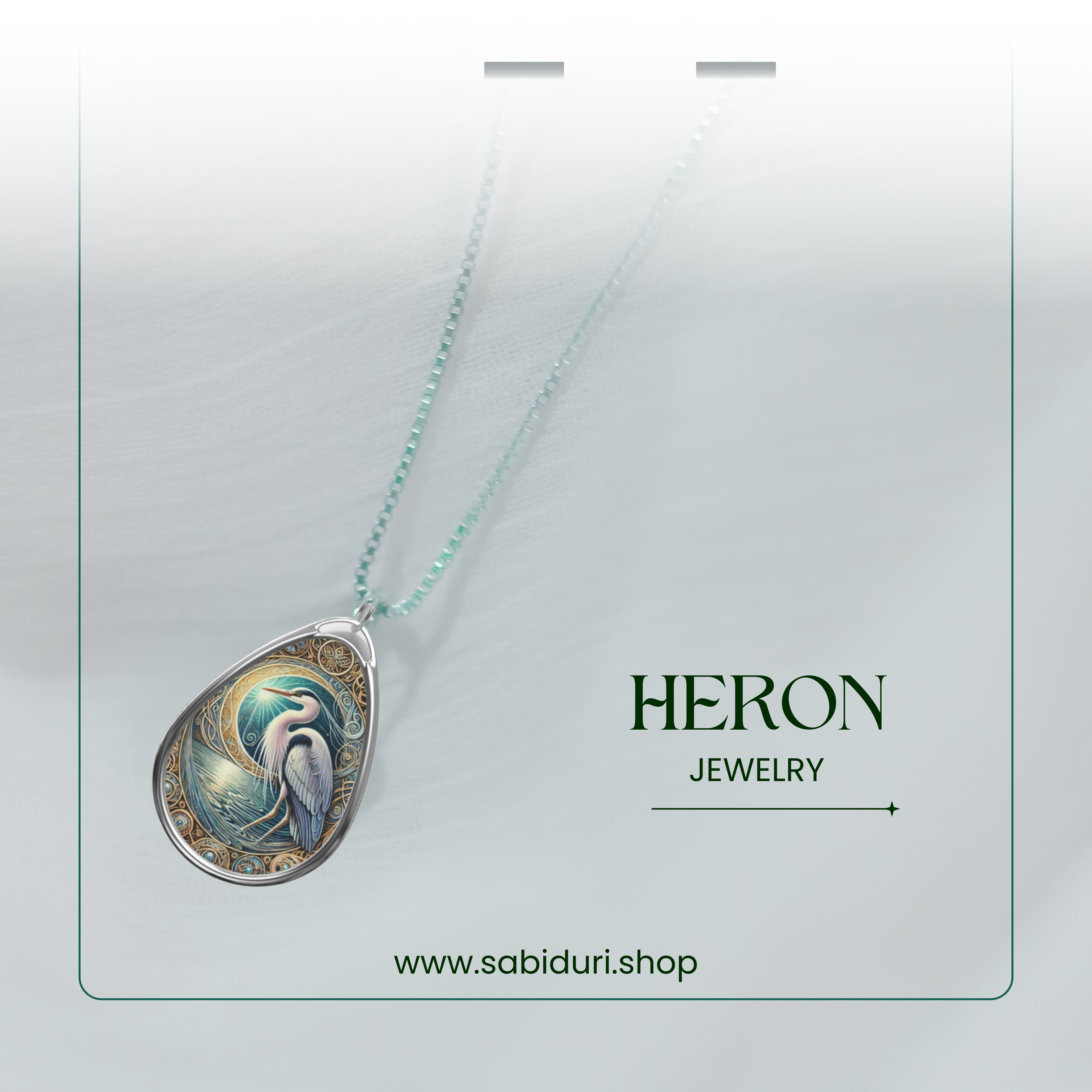 A silver heron pendant necklace laid against a soft, translucent white fabric backdrop. The pendant showcases a detailed heron by water, encased in a gold and blue design. The words "HERON JEWELRY" and "www.sabiduri.shop" are subtly included.