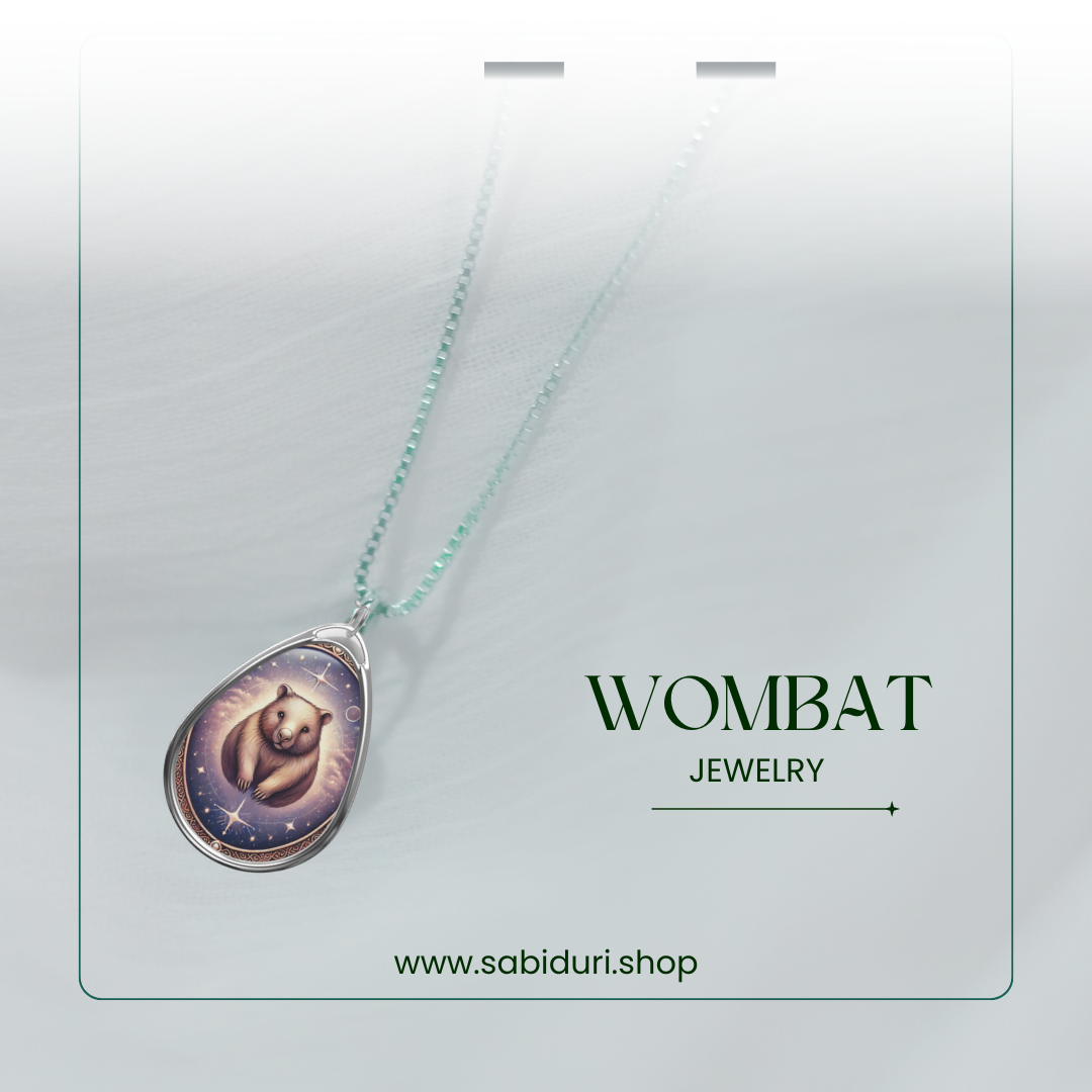 A glowing silver pendant with a celestial wombat illustration, suspended from a dainty beaded chain. The jewelry is presented on a soft, white gradient background with "Wombat Jewelry" and "www.sabiduri.shop" in elegant green text.