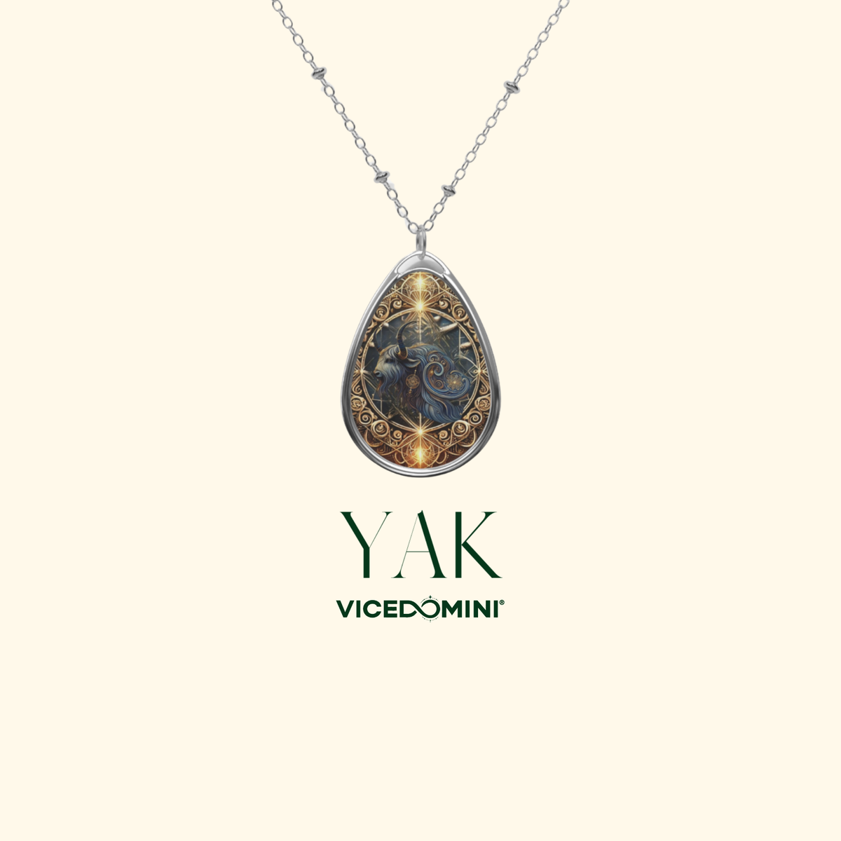 A teardrop-shaped pendant necklace with a silver-toned chain, featuring an ornate fantasy illustration of a blue dragon with gold accents. The brand name 'YAK VICEDOMINI' is displayed below the necklace on a solid pale cream background.