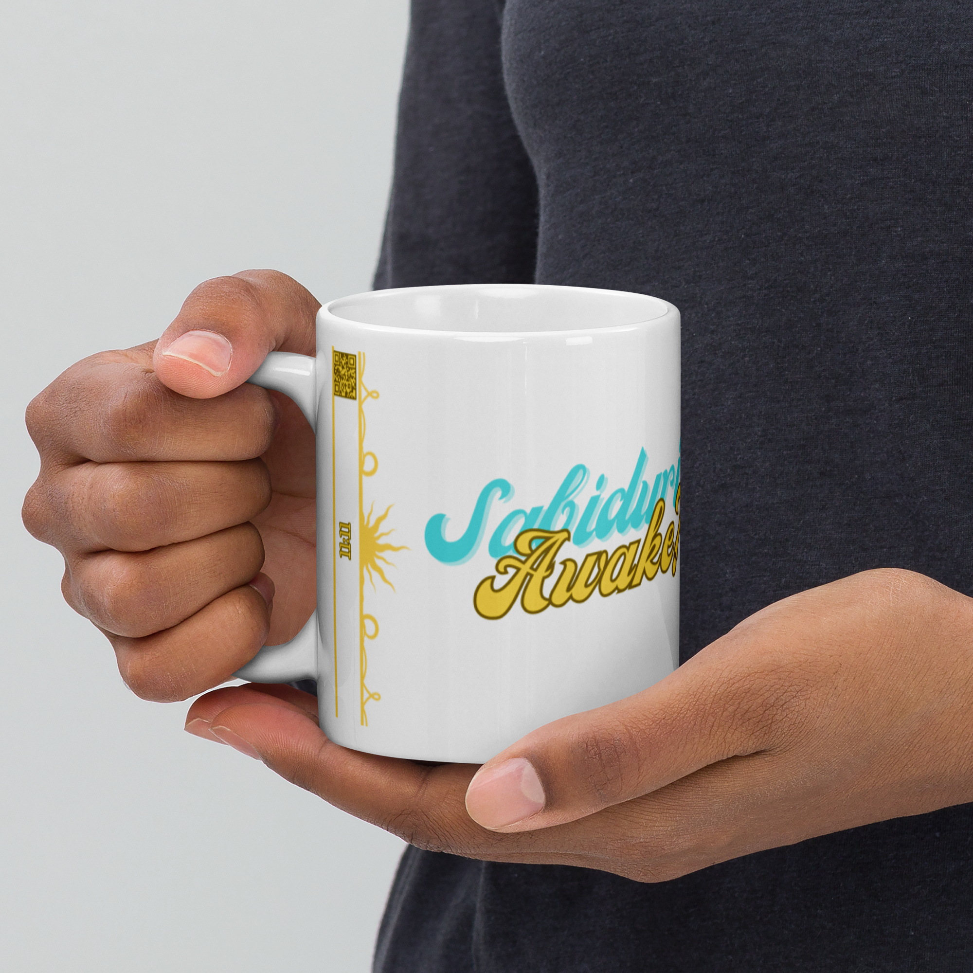 Hands holding a white ceramic mug featuring the phrase "Sabiduría Awake?" in a bold, playful design. The background is simple, emphasizing the mug's design.